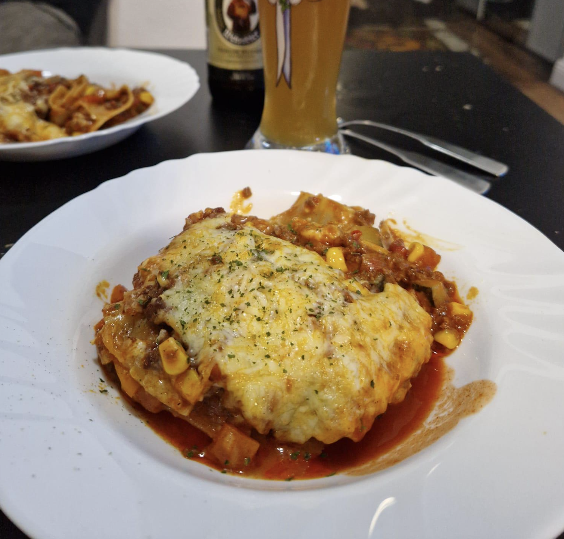 A serving of cheesy baked lasagna sits on a white plate, topped with herbs. In the background, there’s a glass of beer and part of another plate with food visible.