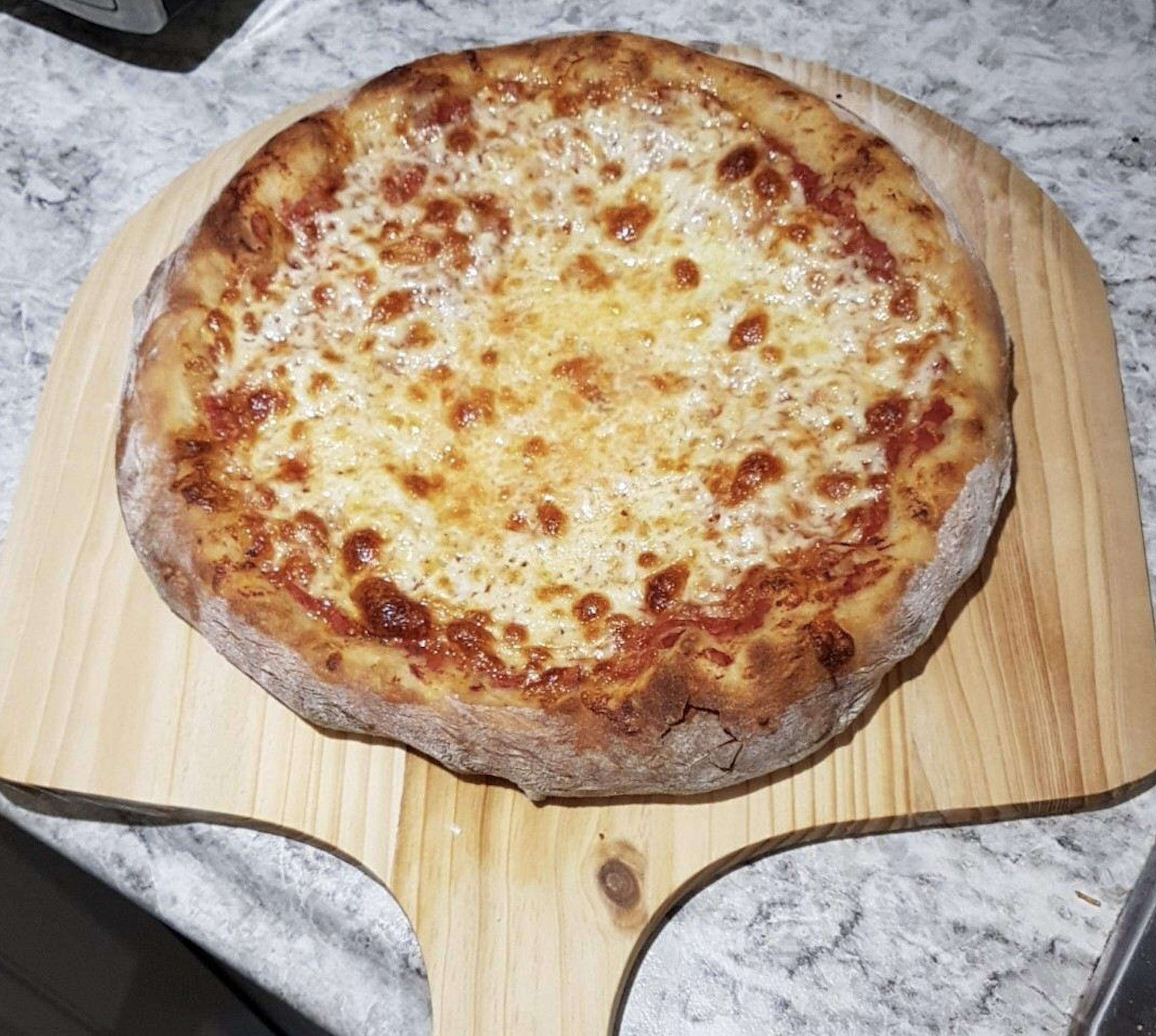 A freshly baked cheese pizza with a golden-brown crust sits on a wooden pizza peel, resting on a speckled granite countertop.