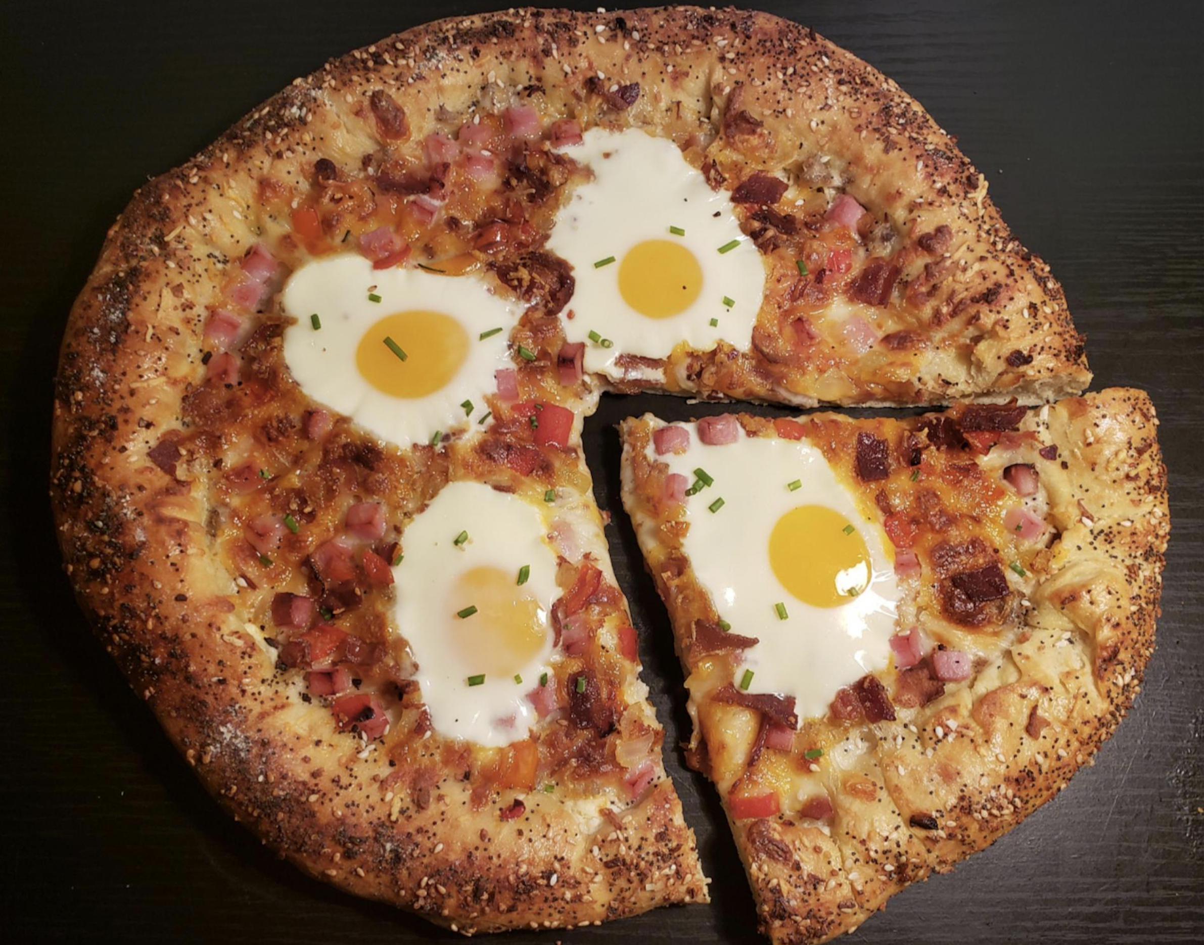 A round pizza with a seeded crust, topped with four sunny-side-up eggs, diced ham, bacon, cheese, and chopped chives; one slice is slightly pulled out from the whole pizza.