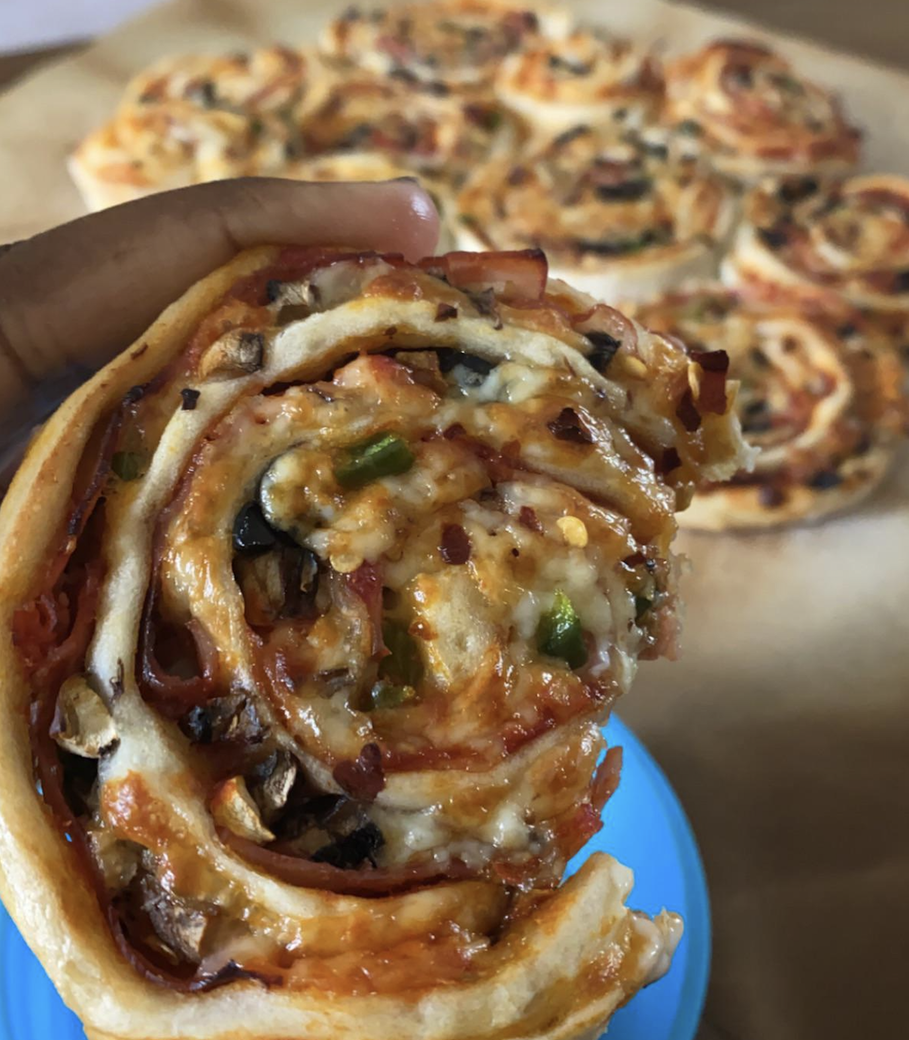 A hand holds a cheesy, spiral pizza roll filled with vegetables and toppings. More pizza rolls are visible on a parchment-lined tray in the background.