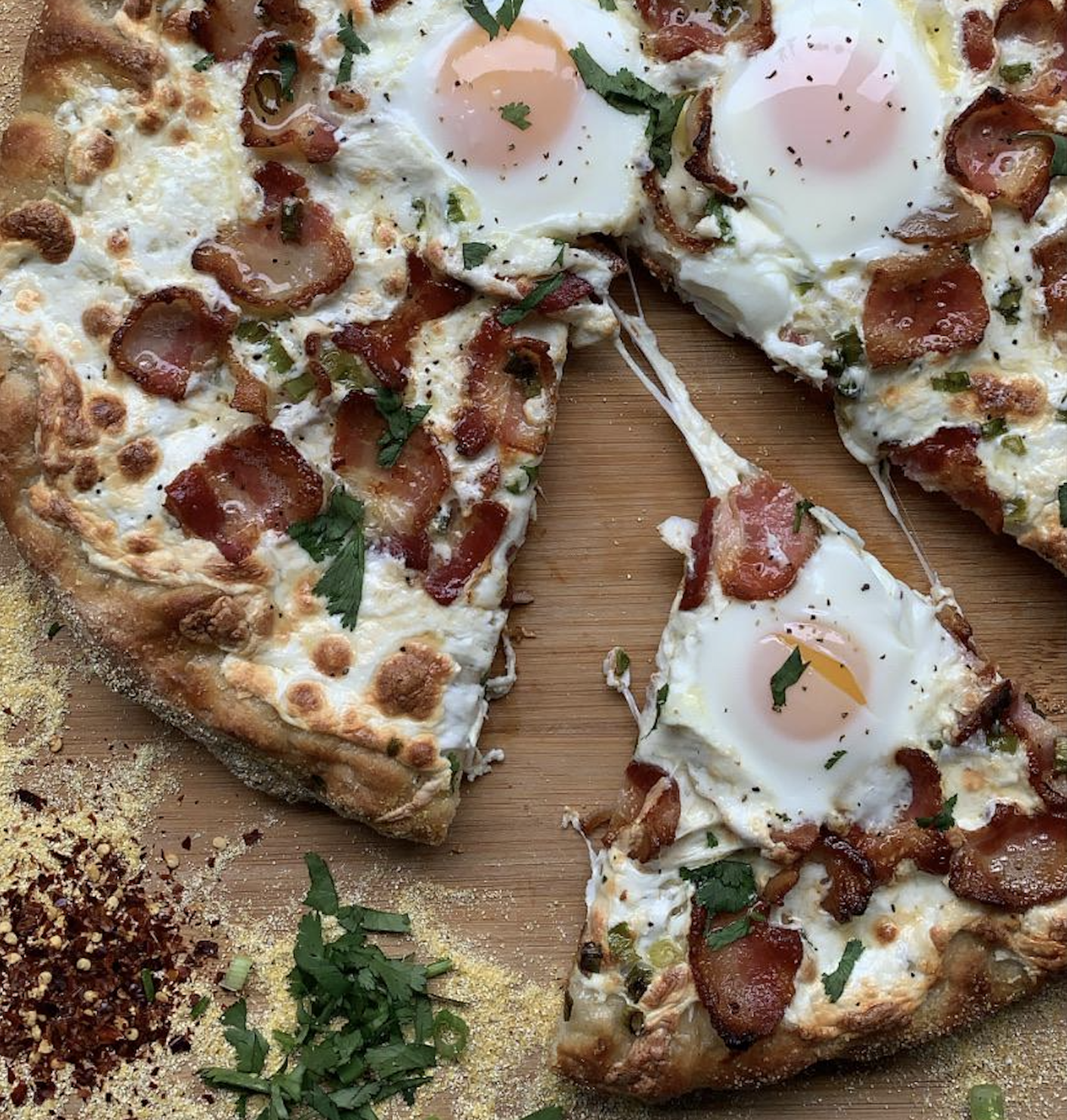 A pizza topped with sunny-side-up eggs, crispy bacon, and fresh herbs sits on a wooden board. One slice is being pulled away, showing melted cheese. Crushed red pepper and chopped herbs are scattered nearby.