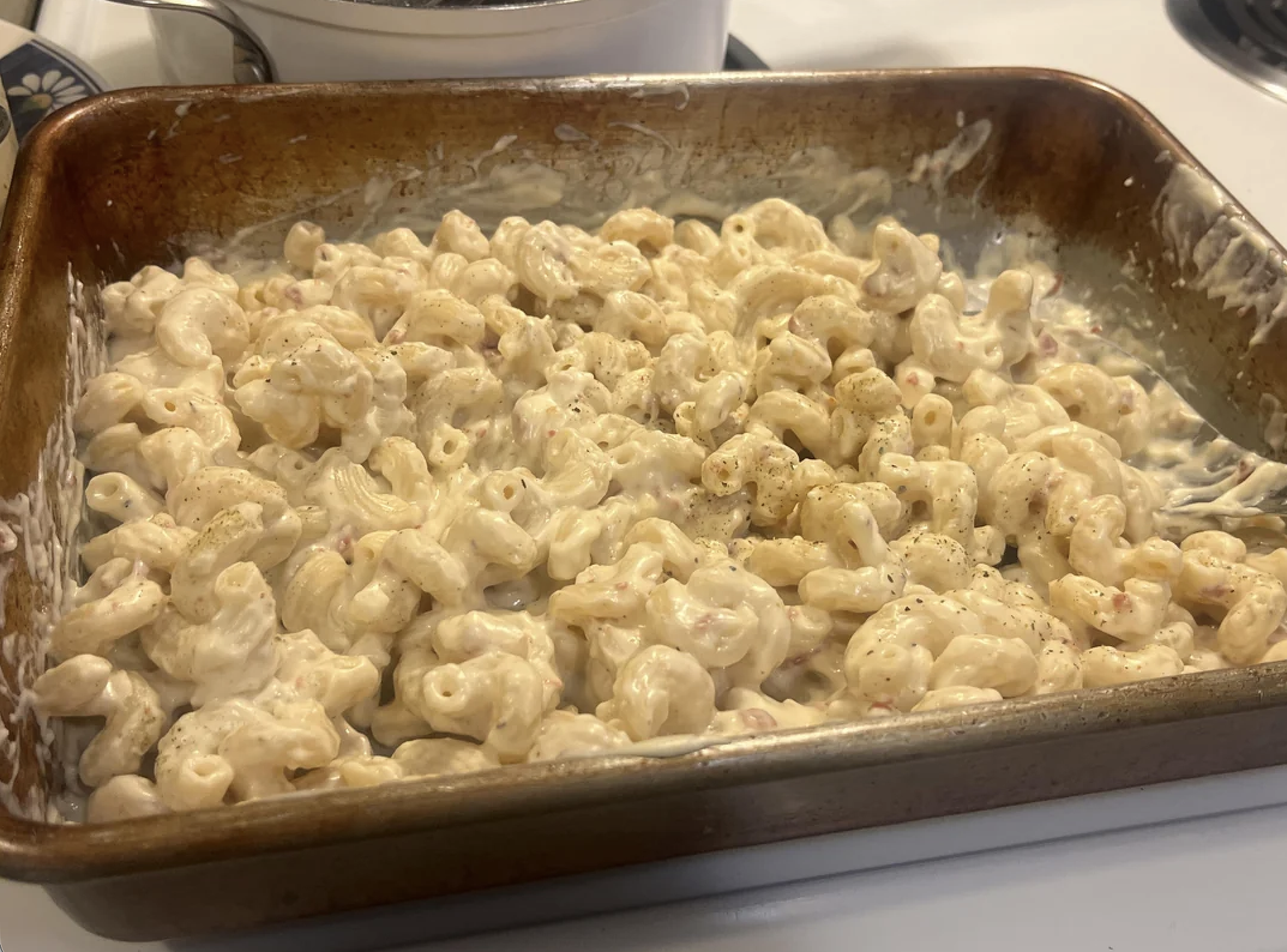 A baking pan filled with creamy macaroni and cheese sits on a stovetop. The pasta is coated in a white cheese sauce and sprinkled with black pepper.