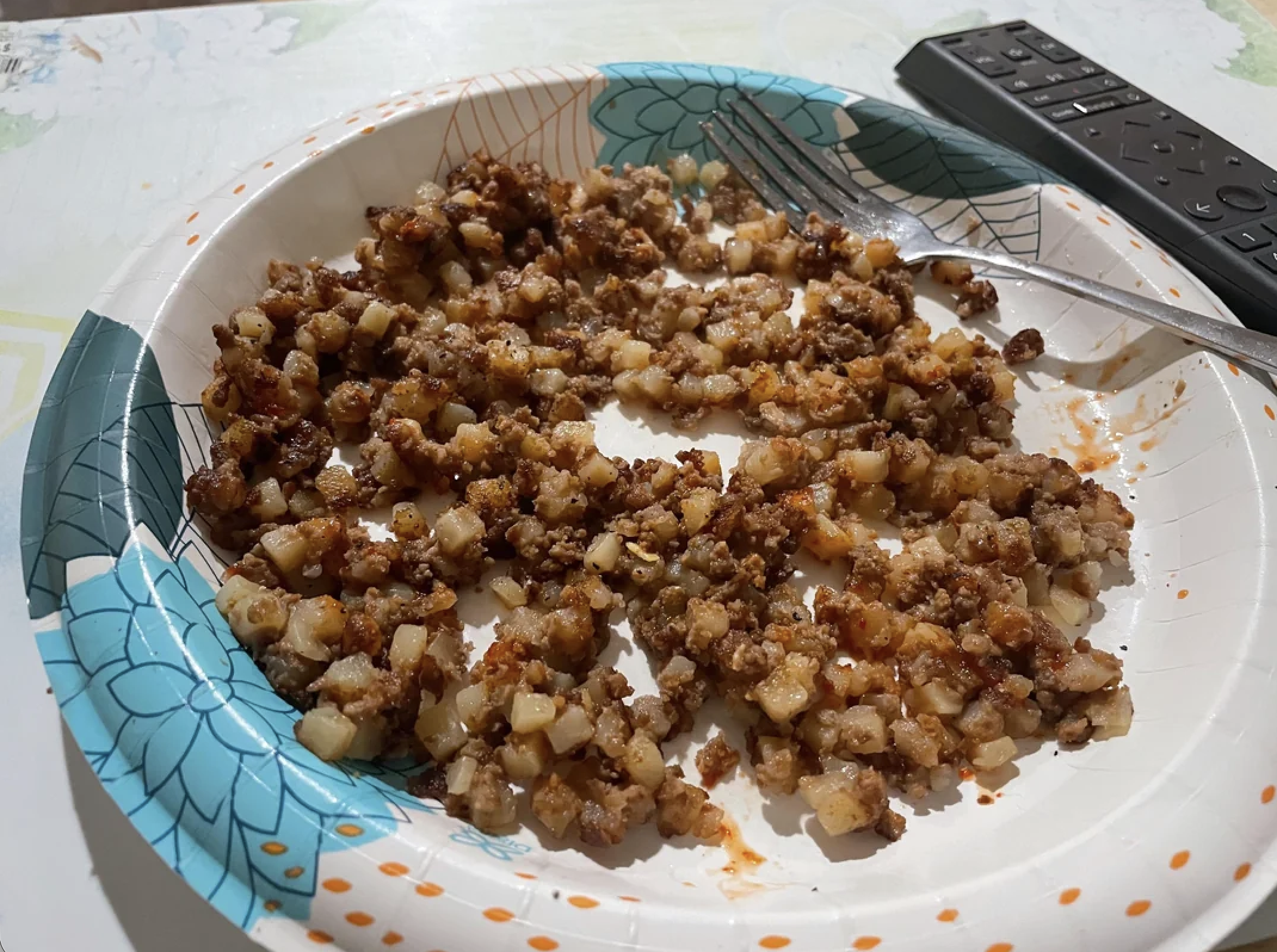 A paper plate with a cooked diced potato and ground meat mixture, a fork, and a TV remote in the background.