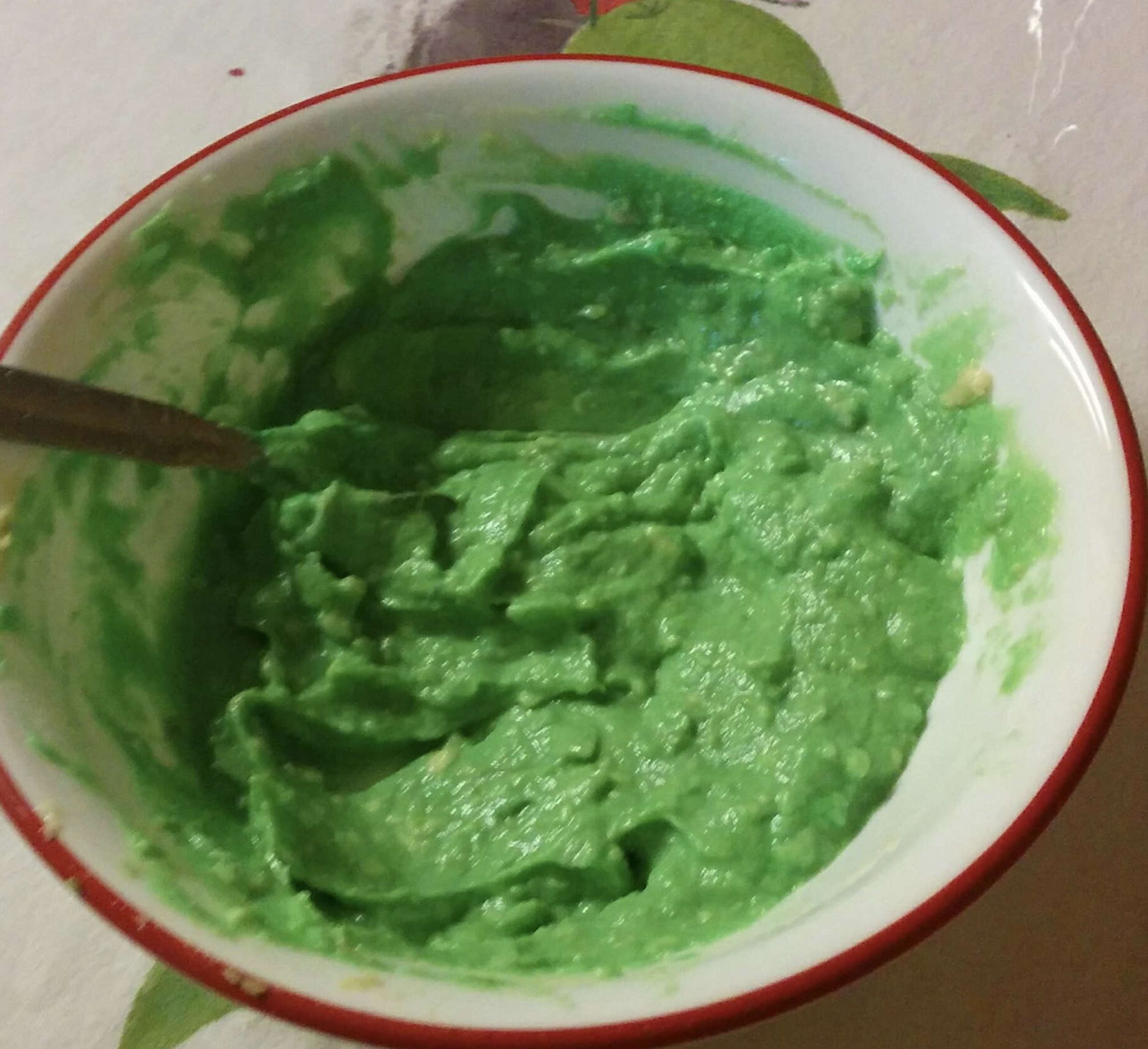 A white bowl with a red rim holds bright green, mashed guacamole. A silver spoon rests partially submerged in the mixture, which sits on a white table covered with a light-colored tablecloth patterned with green leaves.