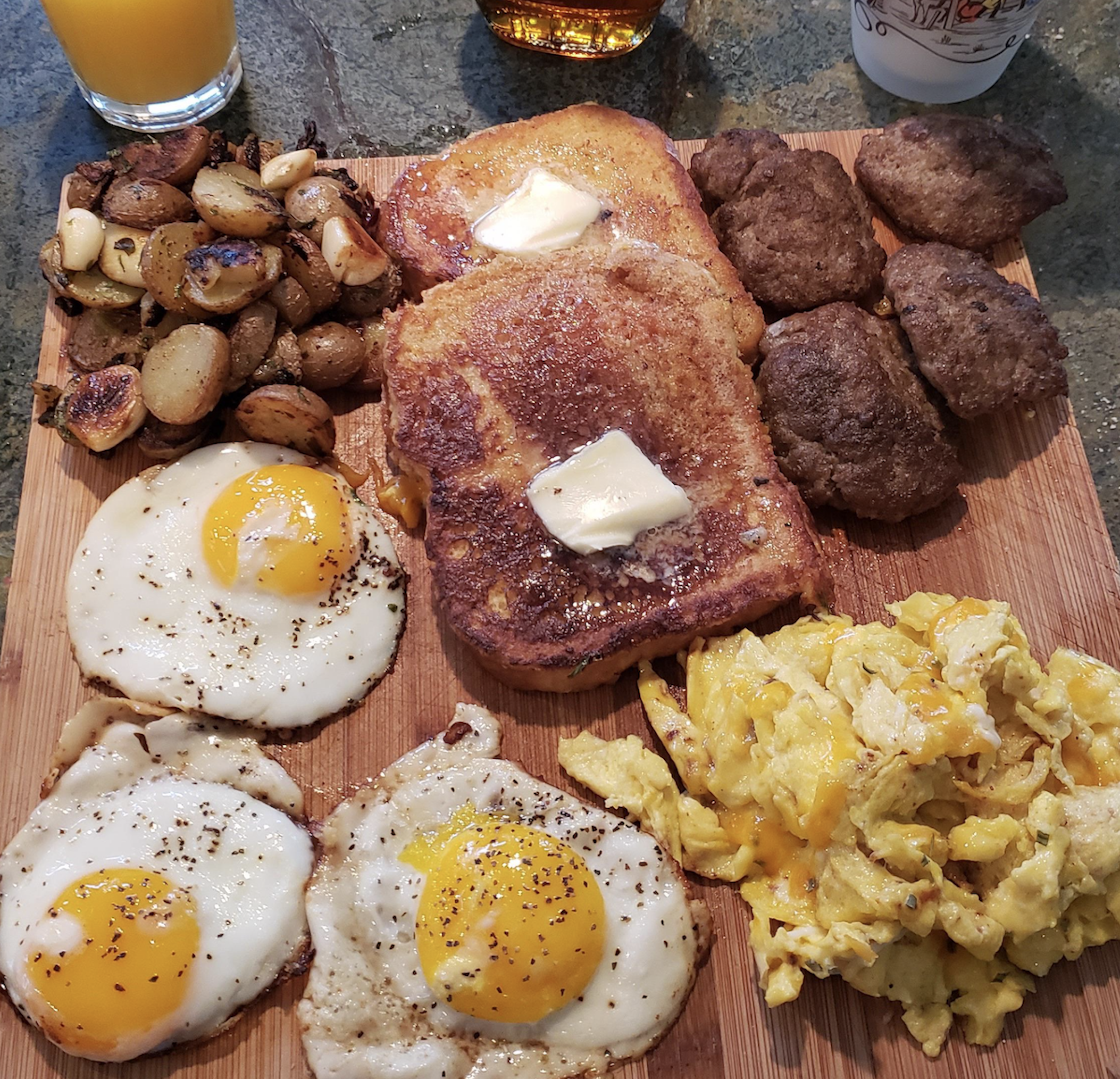 A wooden board topped with a breakfast feast: French toast with melting butter, four sausage patties, sautéed potatoes with garlic, three sunny-side-up eggs, and a large mound of cheesy scrambled eggs, with juice in the background.
