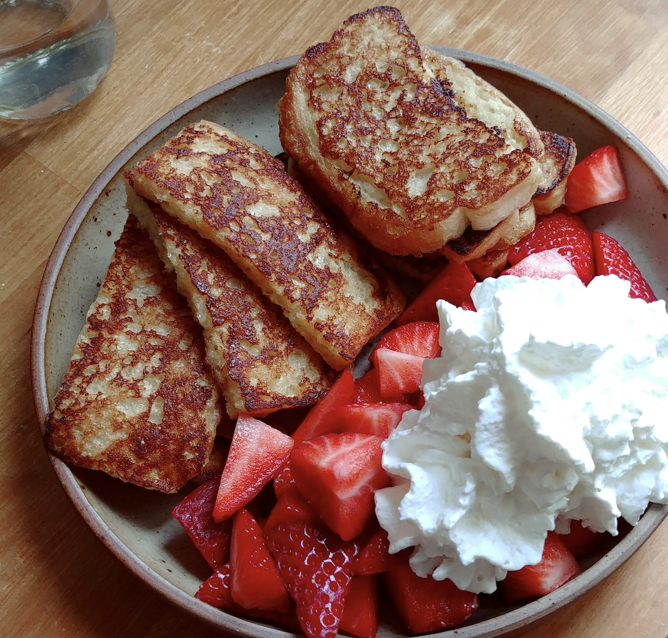 A plate of golden-brown French toast sticks served with sliced fresh strawberries and a large dollop of whipped cream, set on a wooden table.