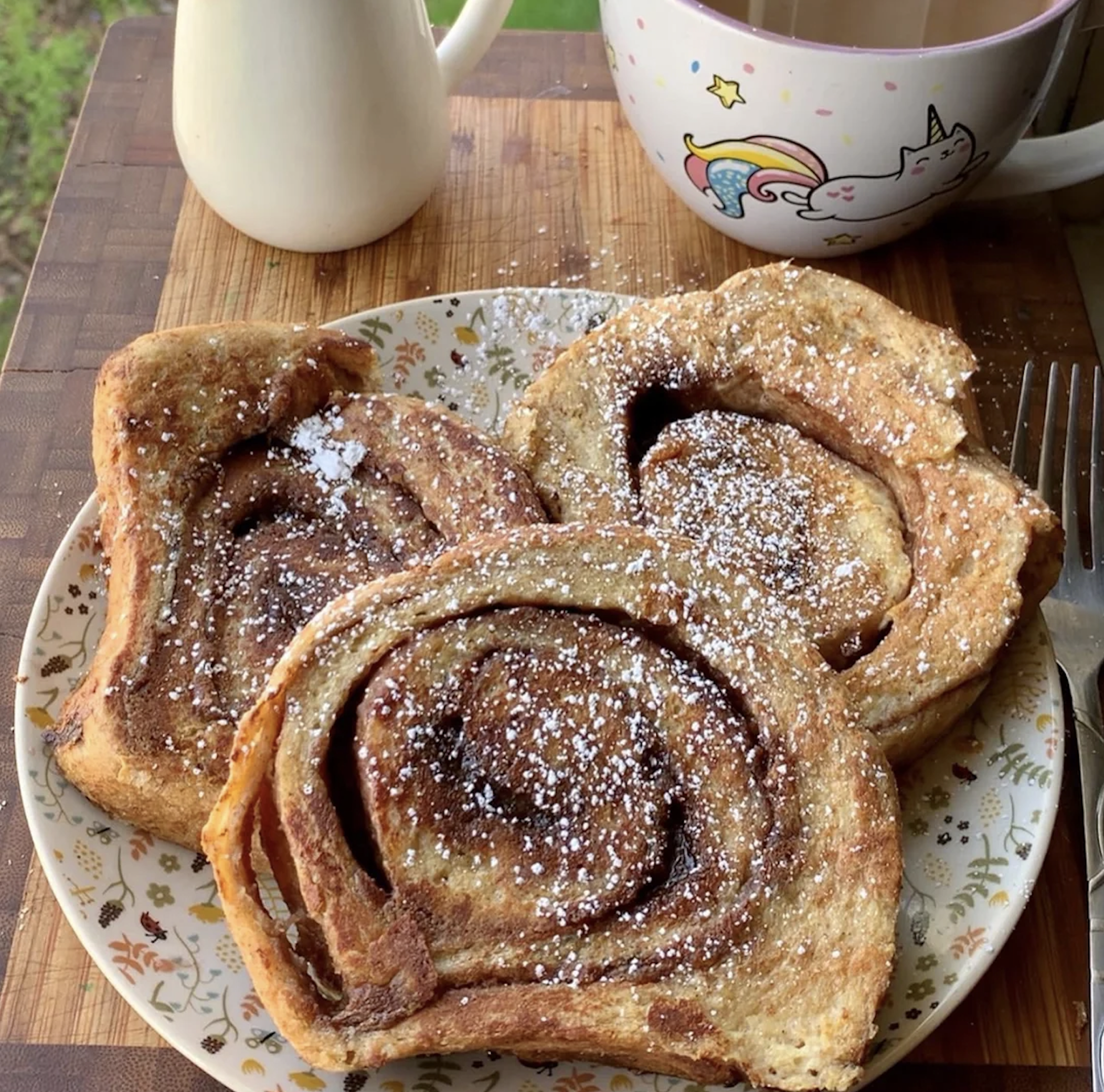 Three slices of golden-brown cinnamon swirl French toast, dusted with powdered sugar, sit on a floral plate. A fork rests to the side, with a unicorn mug and small pitcher blurred in the background on a wooden surface.