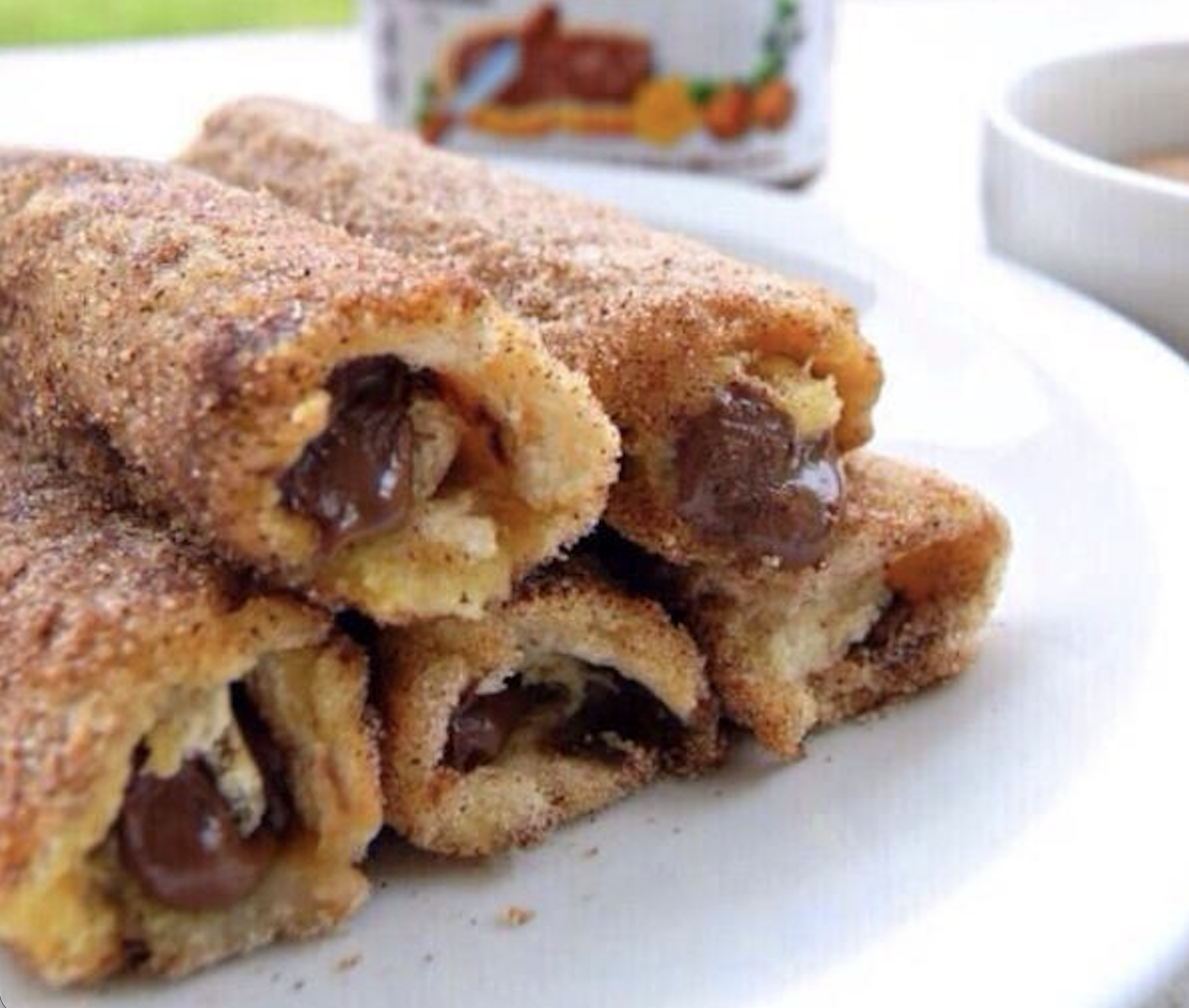 Five cinnamon sugar-coated rolled toast sticks filled with chocolate hazelnut spread are stacked on a white plate, with blurred jars and bowls in the background.