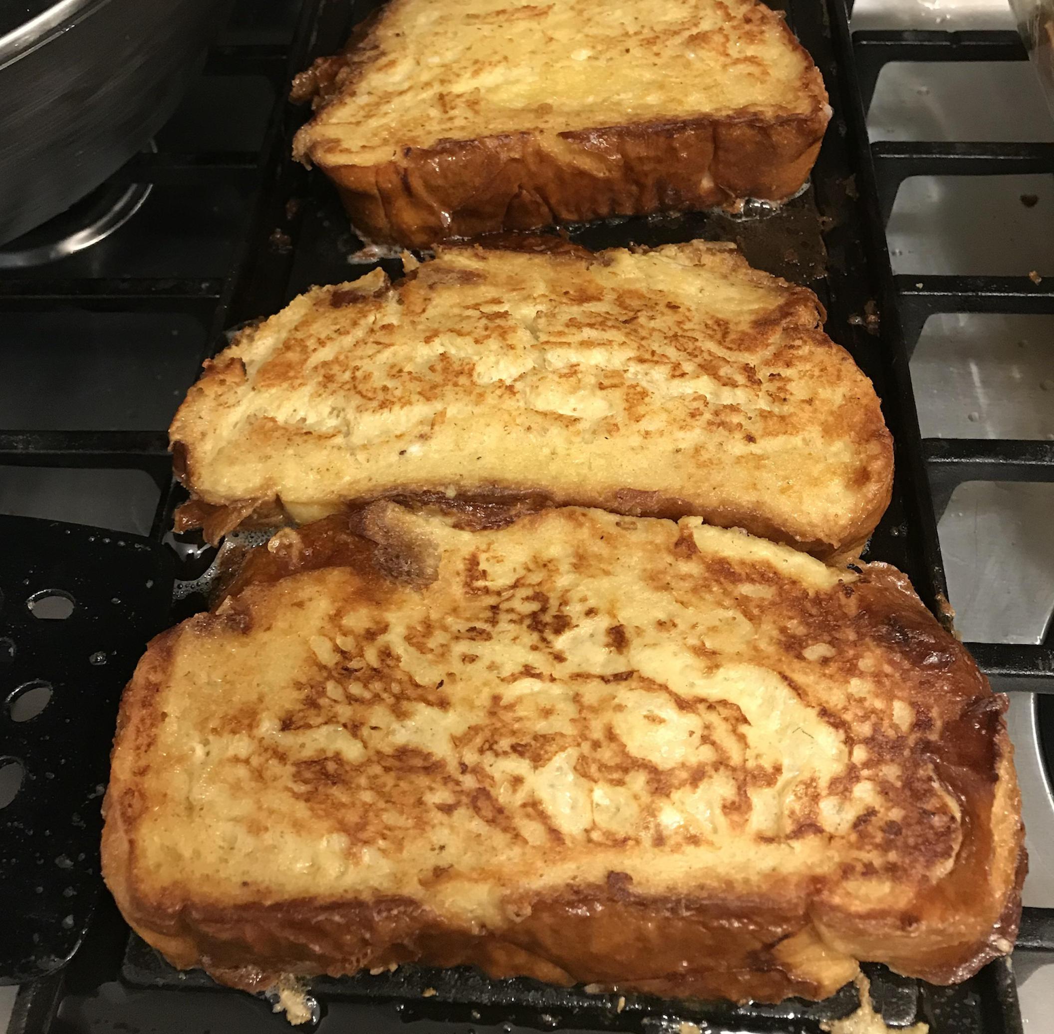 Three slices of golden-brown French toast cooking on a black griddle sitting on a stovetop.