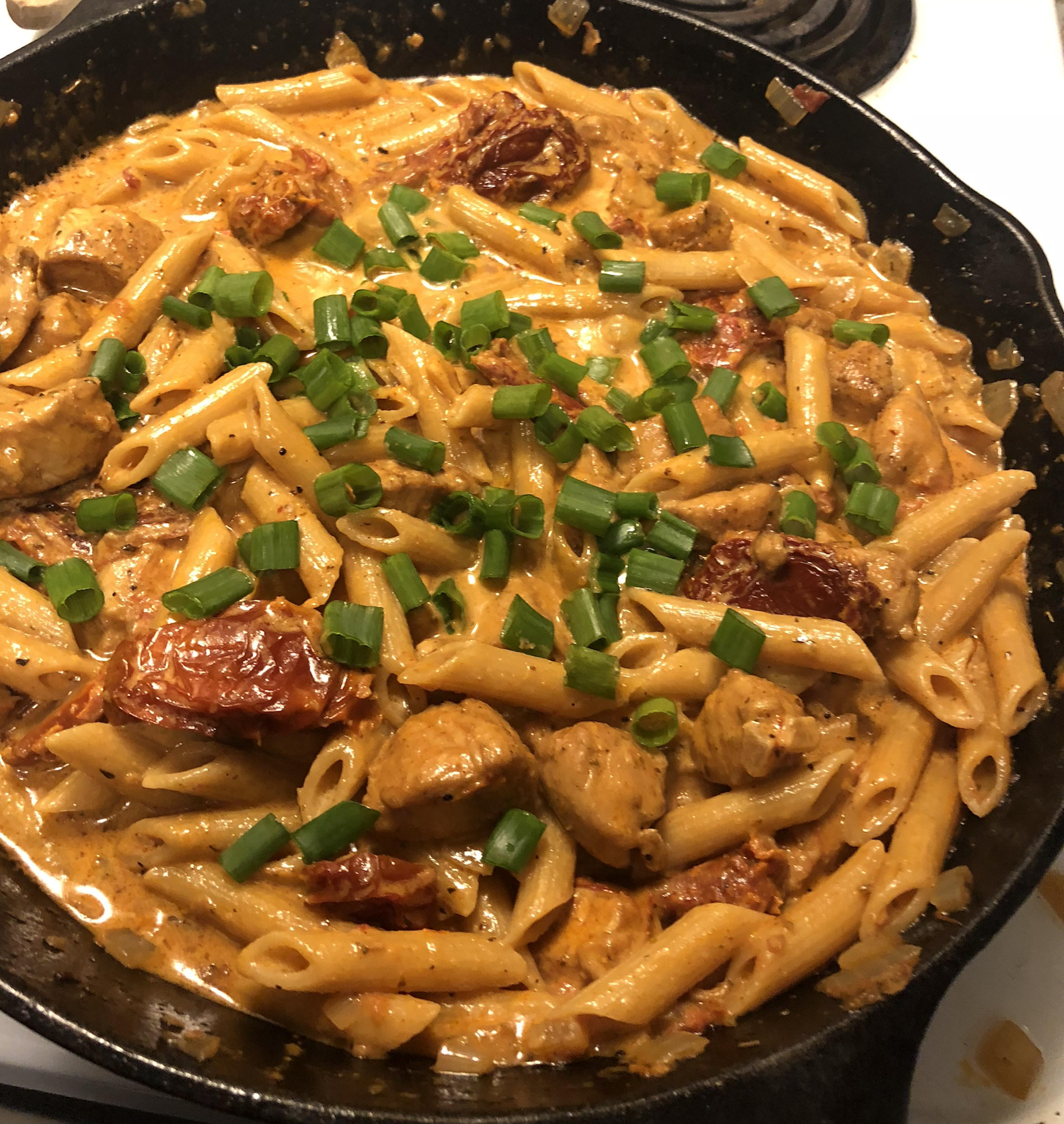 Penne pasta in a creamy sauce with chunks of chicken, sun-dried tomatoes, and sliced green onions, served in a black skillet on a stove.