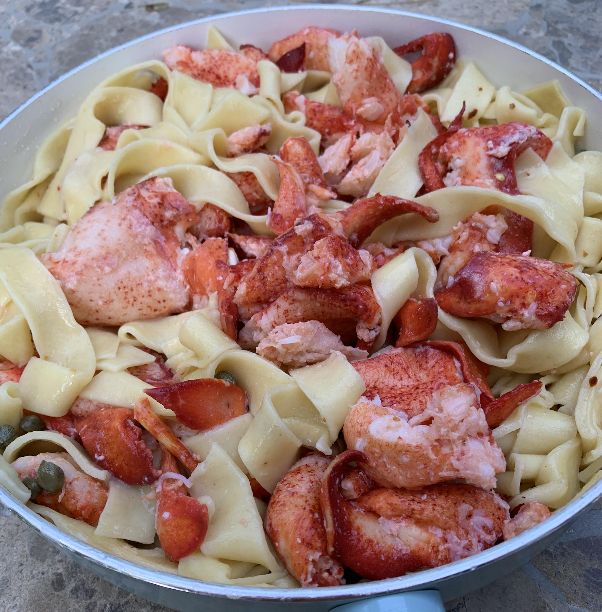 A pan filled with wide pappardelle pasta and large chunks of lobster meat on a stone surface.