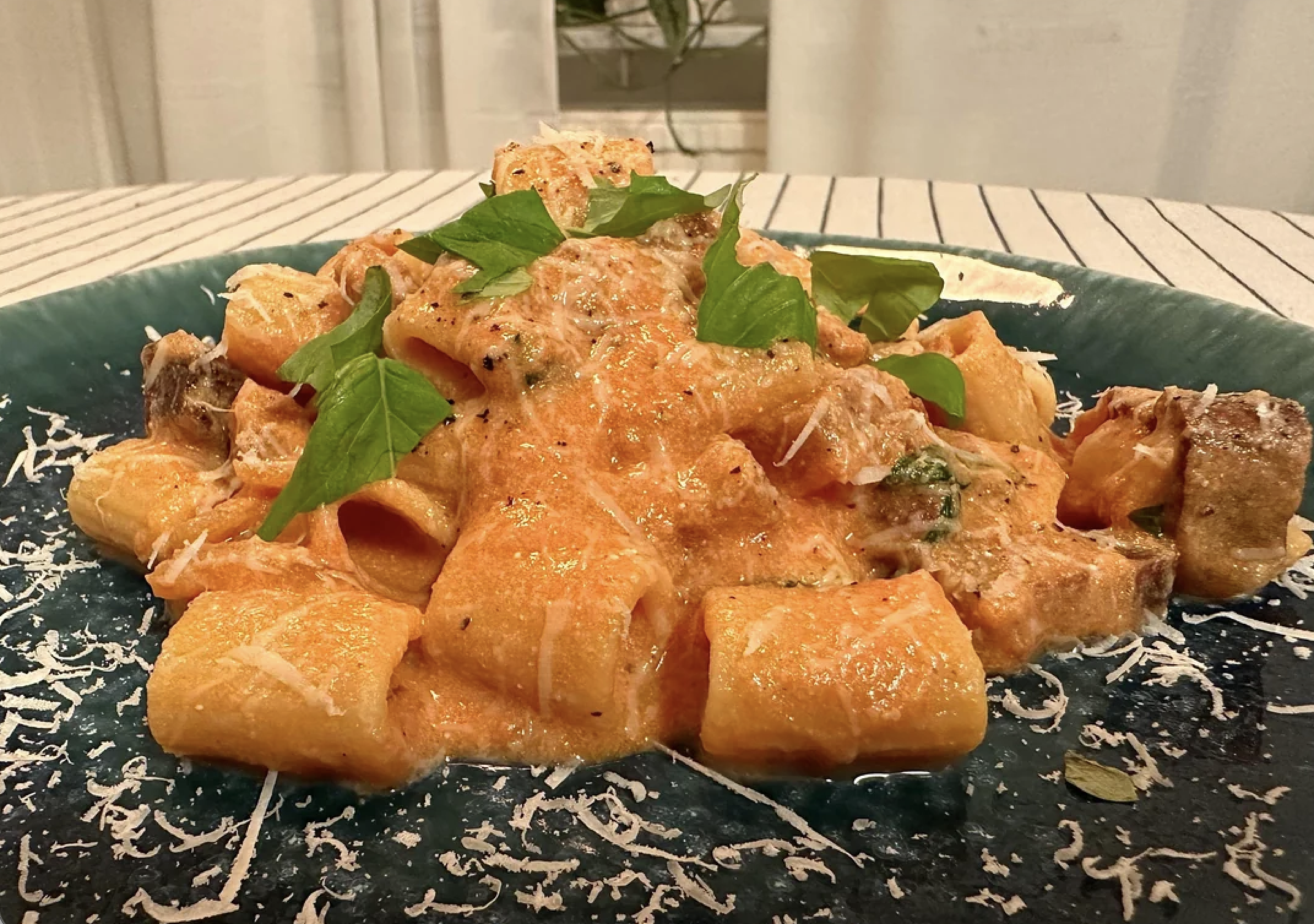 A plate of rigatoni pasta in creamy tomato sauce, topped with grated cheese and fresh basil leaves, served on a dark green dish with a striped tablecloth underneath.