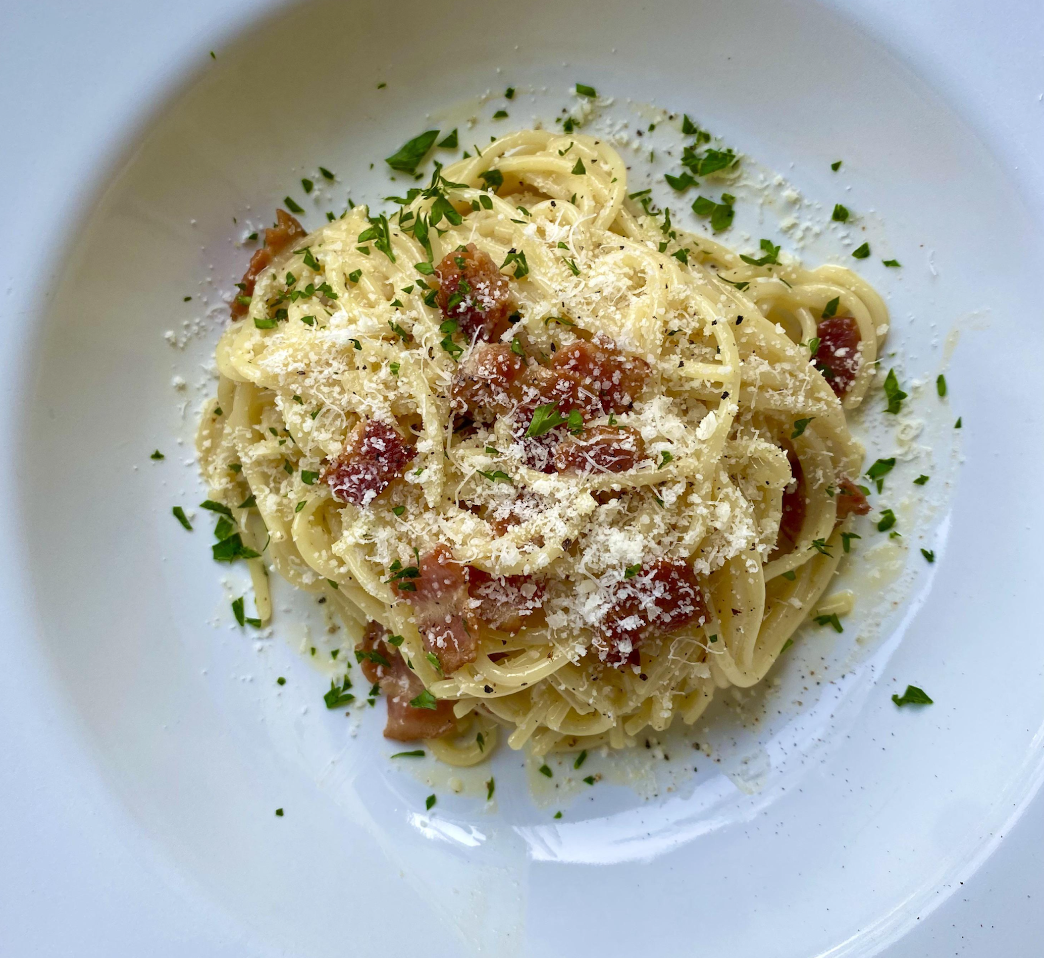 A plate of spaghetti carbonara topped with grated cheese, chopped parsley, and pieces of bacon or pancetta, served on a white dish.