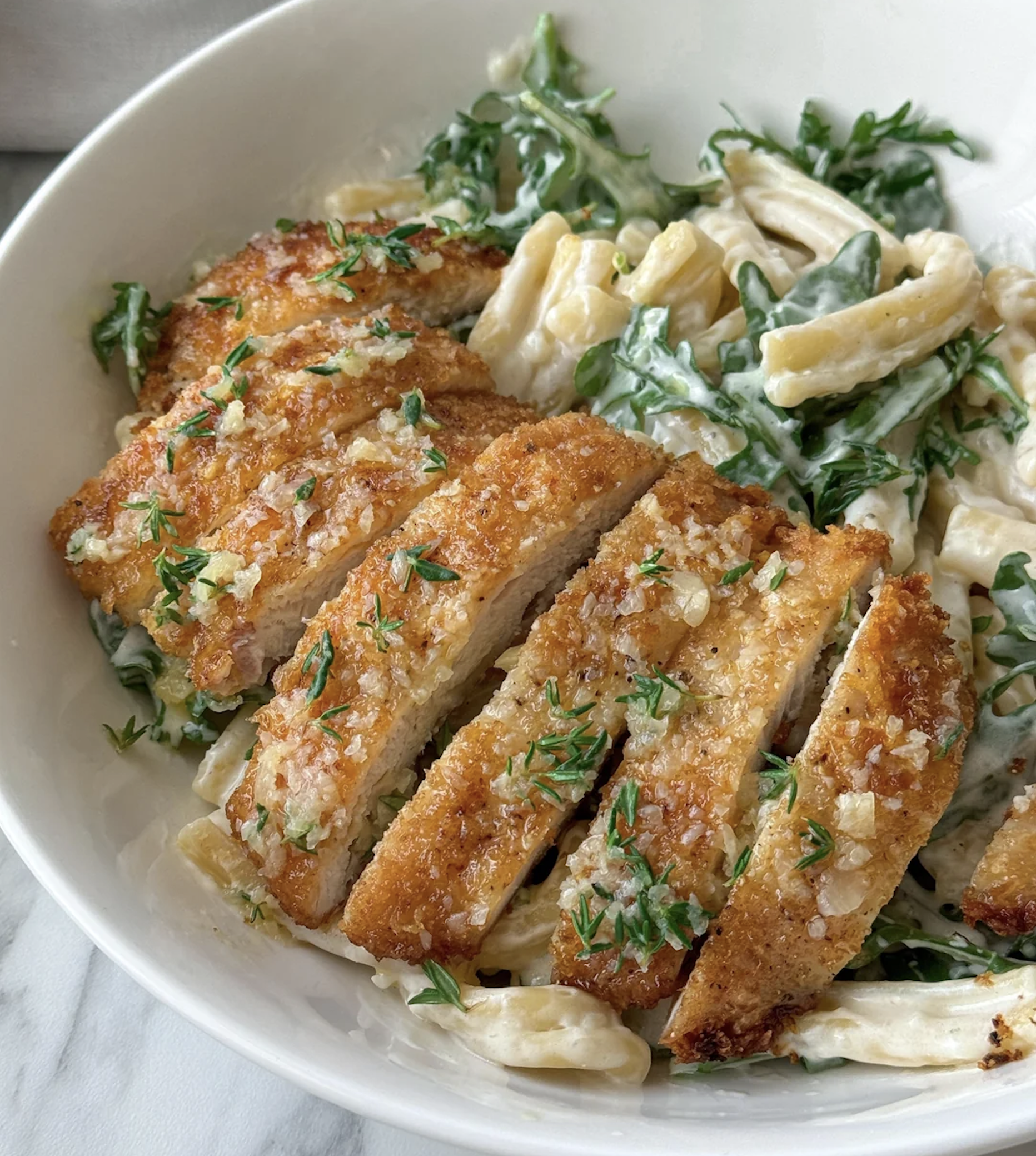 Sliced crispy breaded chicken breast on creamy pasta with spinach, garnished with chopped herbs, served in a white bowl on a marble surface.