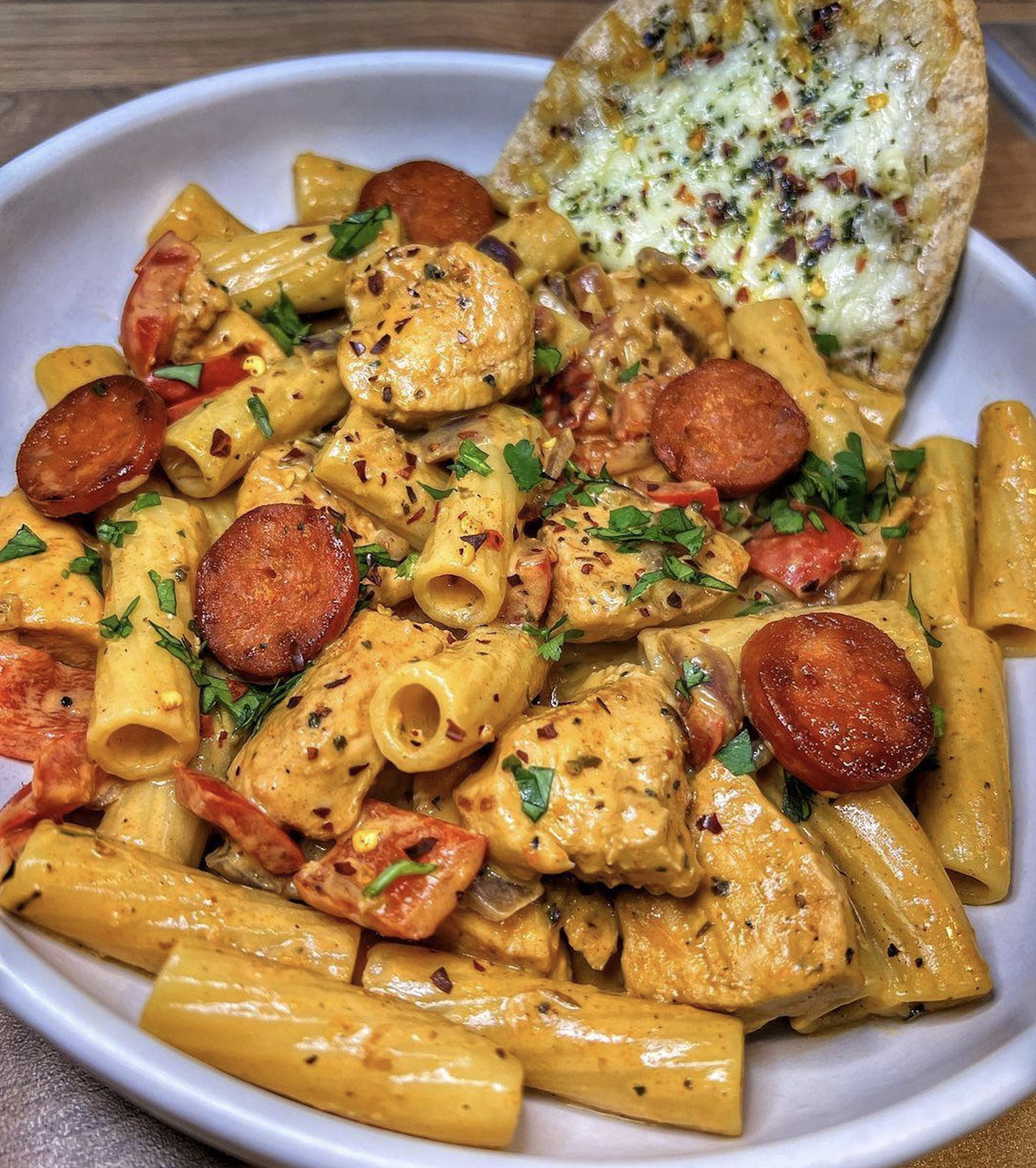 A bowl of creamy rigatoni pasta with grilled chicken, sliced sausage, red bell peppers, fresh herbs, and a piece of garlic bread topped with herbs.