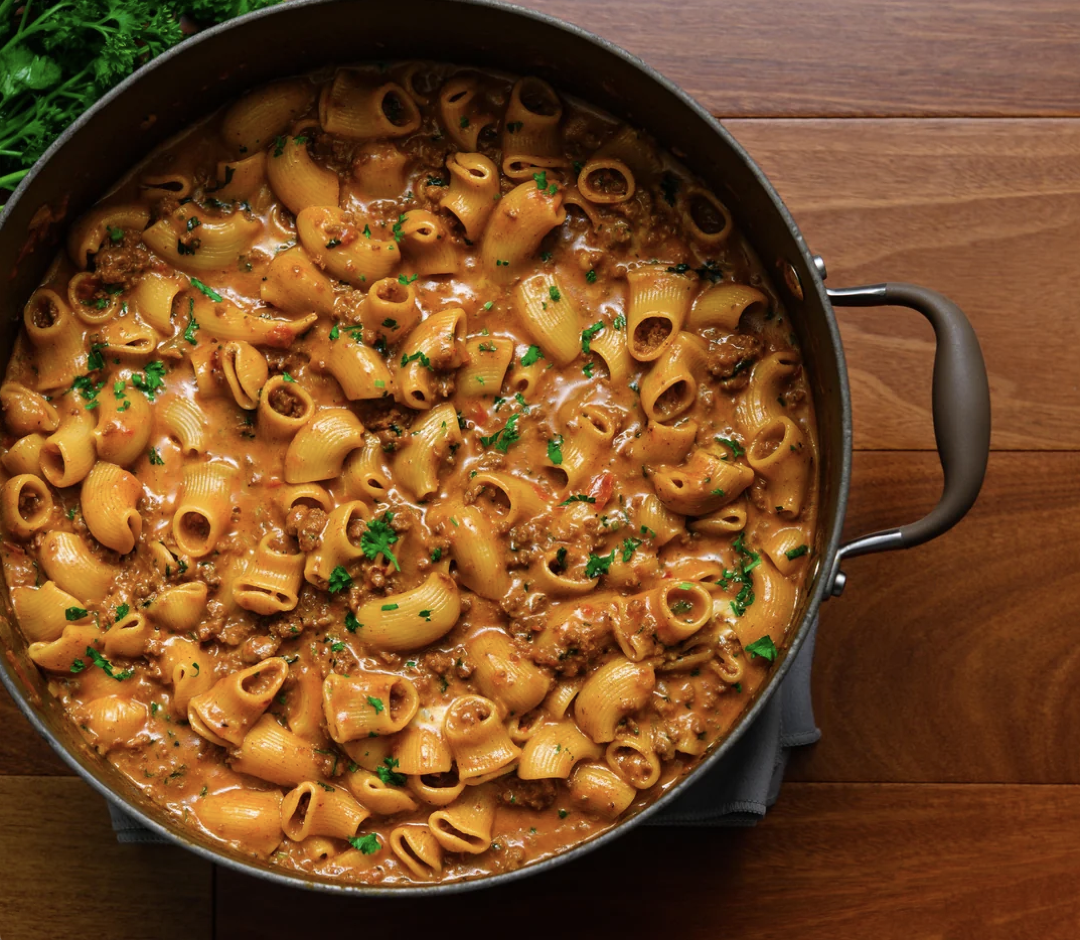 A pot of creamy pasta with tube-shaped noodles, ground meat, and a tomato-based sauce, garnished with chopped parsley, sits on a wooden surface.