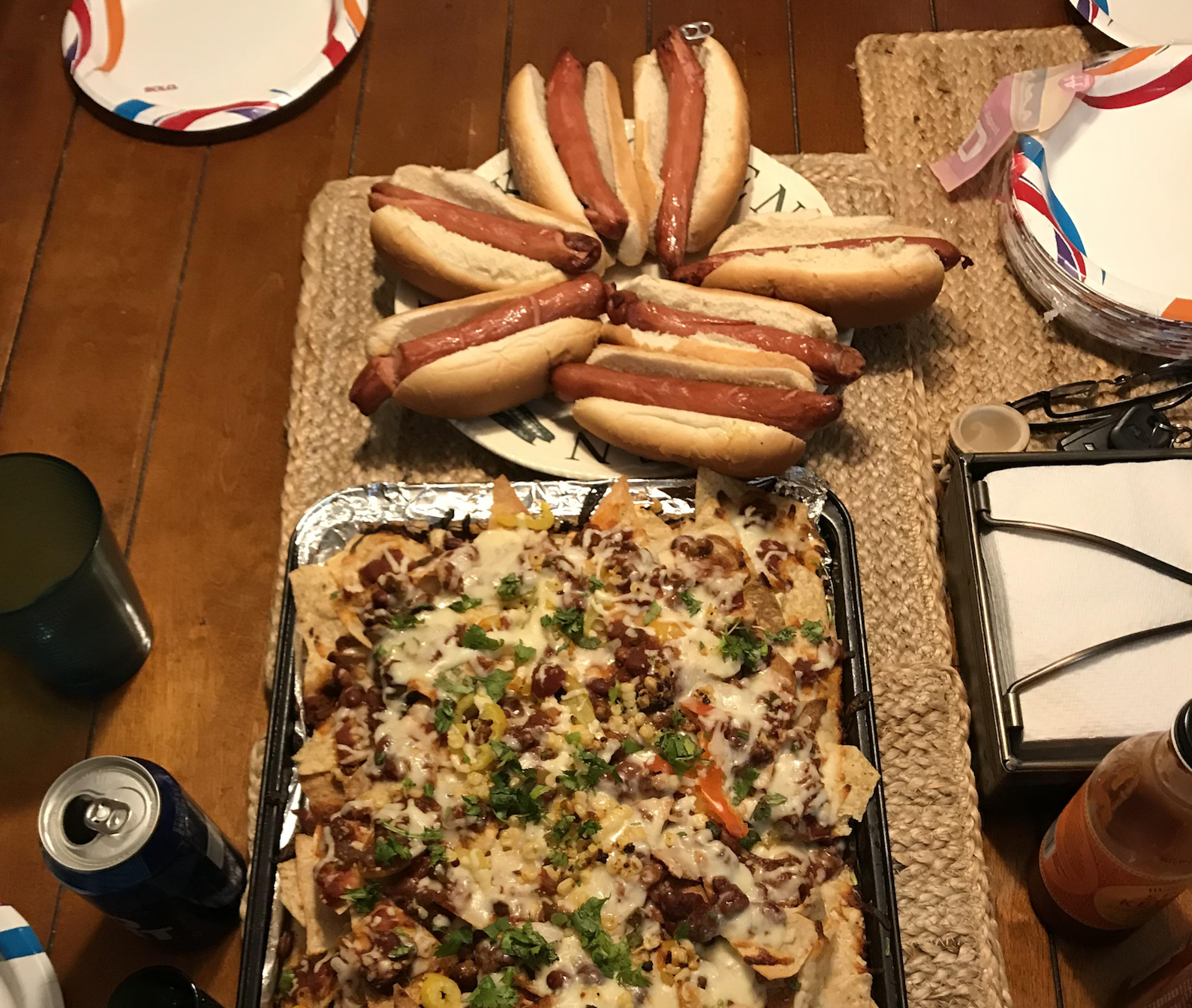 A tray of loaded nachos with melted cheese and toppings sits in front of a plate stacked with six hot dogs in buns on a table set for a meal.