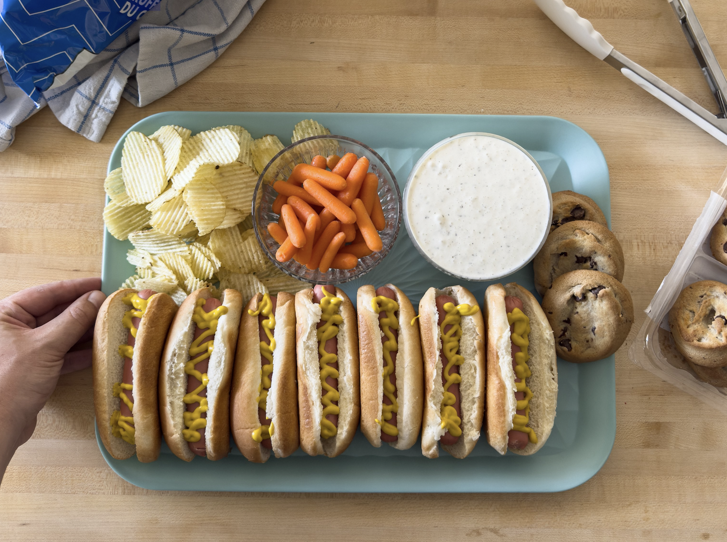 A tray with six hot dogs topped with mustard, ridged potato chips, baby carrots, ranch dip, and chocolate chip cookies. A hand holds the tray on a wooden table with kitchen items nearby.