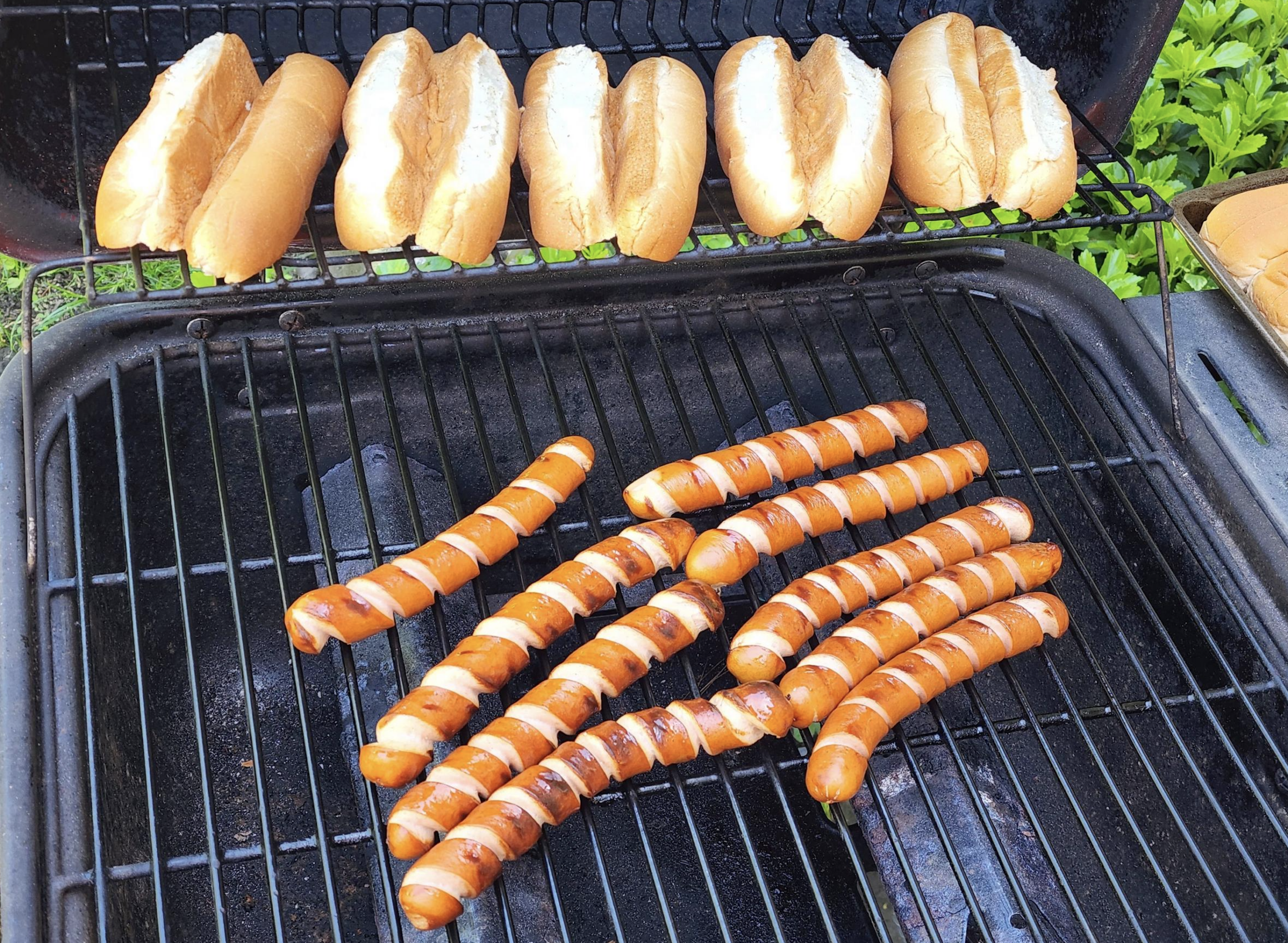 Hot dog buns are on the top rack of a grill, while several grilled hot dogs with char marks are cooking on the bottom grill rack. Greenery is visible in the background.