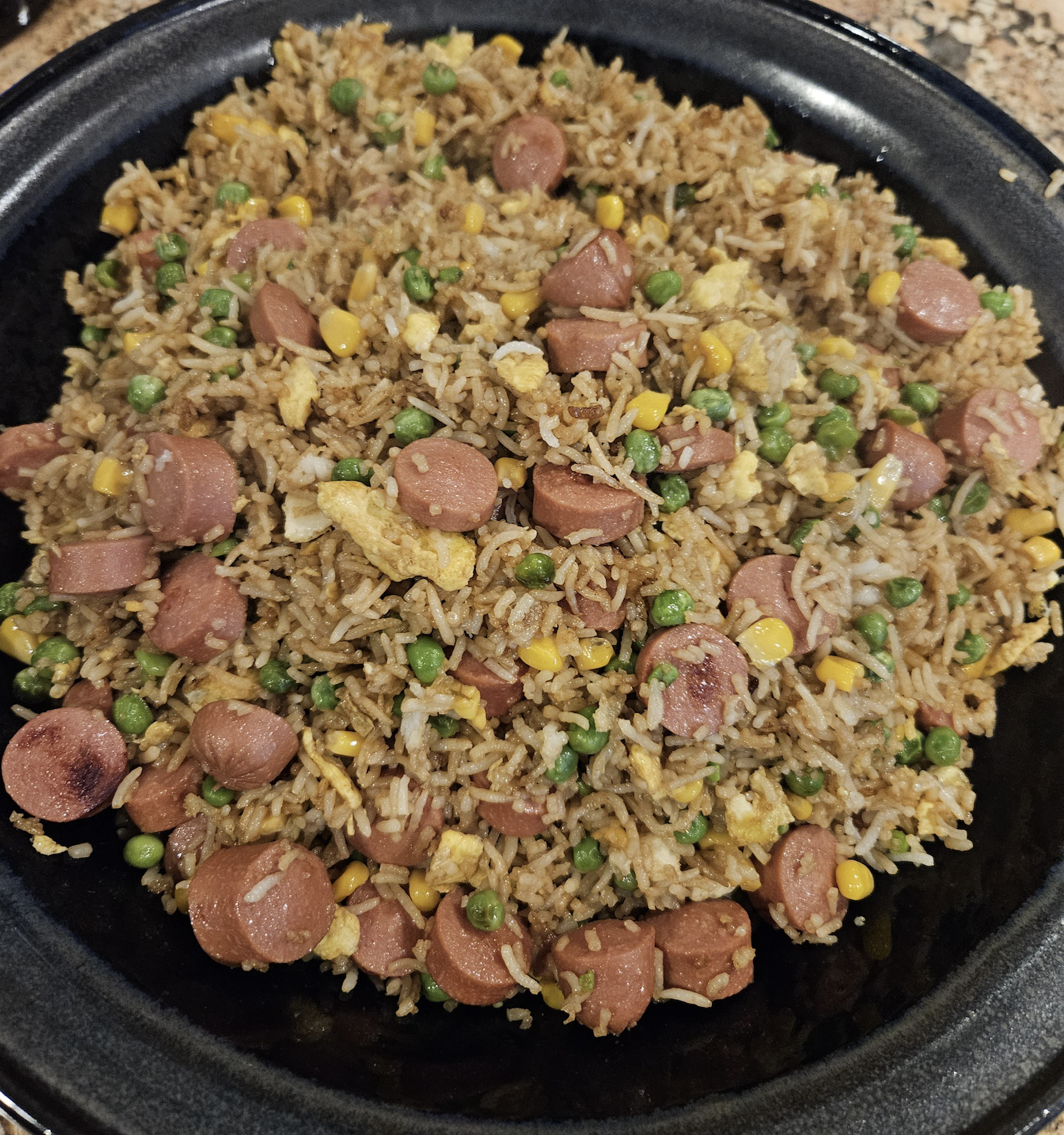A plate of fried rice mixed with sliced hot dogs, scrambled eggs, peas, and corn, served on a black dish.