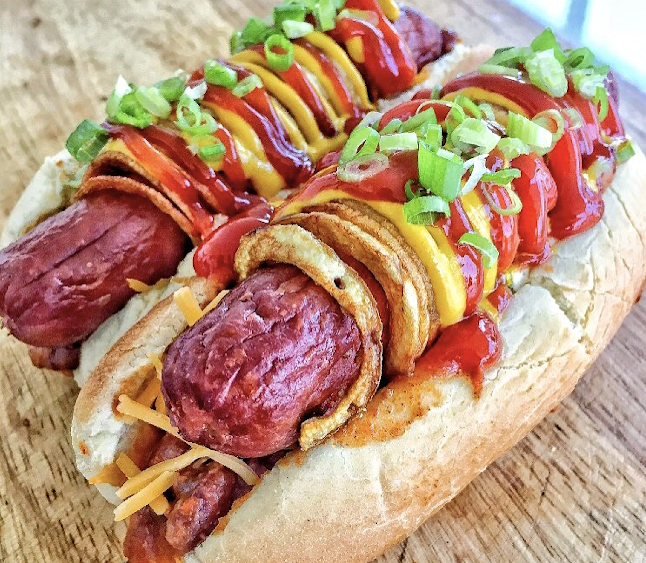 Two hot dogs in buns topped with ketchup, mustard, sliced green onions, crispy fried onions, and shredded cheese, placed on a wooden surface.
