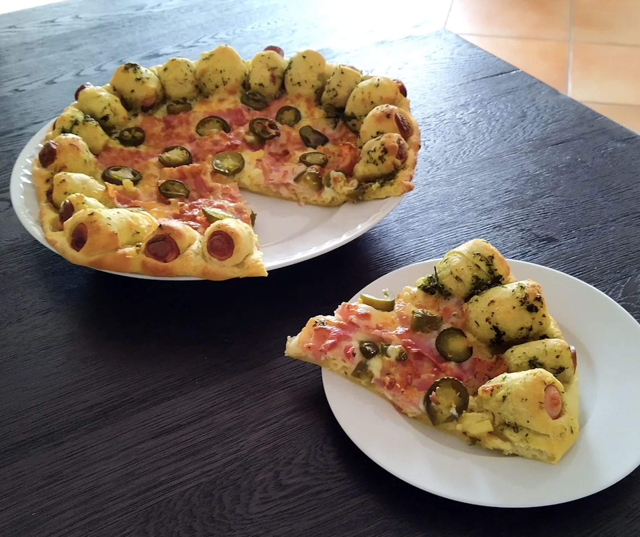 A pizza with a crust made of dough balls filled with sausage, topped with jalapenos, sits on a plate. One slice is served on a smaller plate beside it, on a dark wooden table.