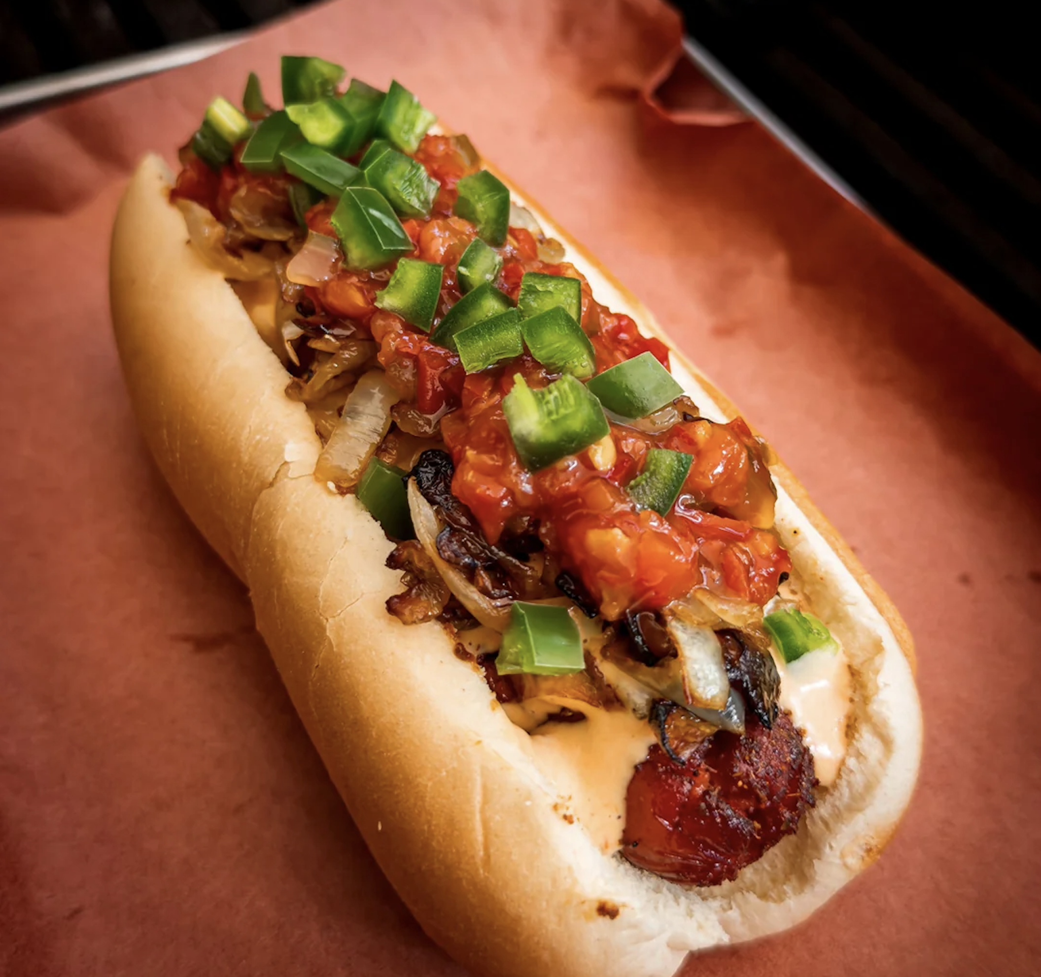 A grilled sausage in a bun topped with chopped onions, diced green peppers, salsa, and sauce, placed on brown paper.