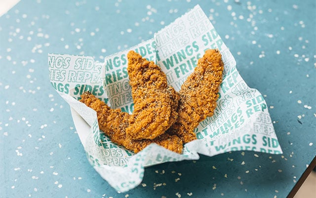 Three crispy fried chicken tenders, reminiscent of the best fried chicken chains, sit on branded paper atop a blue surface sprinkled with salt.