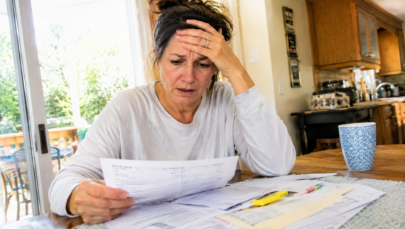 A woman sits at a table, looking worried while reading a bill or financial document&mdash;an all-too-familiar scene for workers struggling financially. Her hand rests on her forehead in concern, surrounded by various papers, a pen, and a mug scattered across the table.