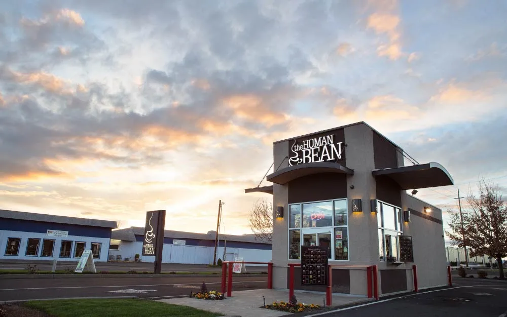 A drive-thru coffee shop with "The Human Bean" sign stands under a dramatic cloudy sky at sunrise or sunset. Surrounded by pavement, this spot blends the cozy charm of coffee houses with the convenience of a menu board and order window.