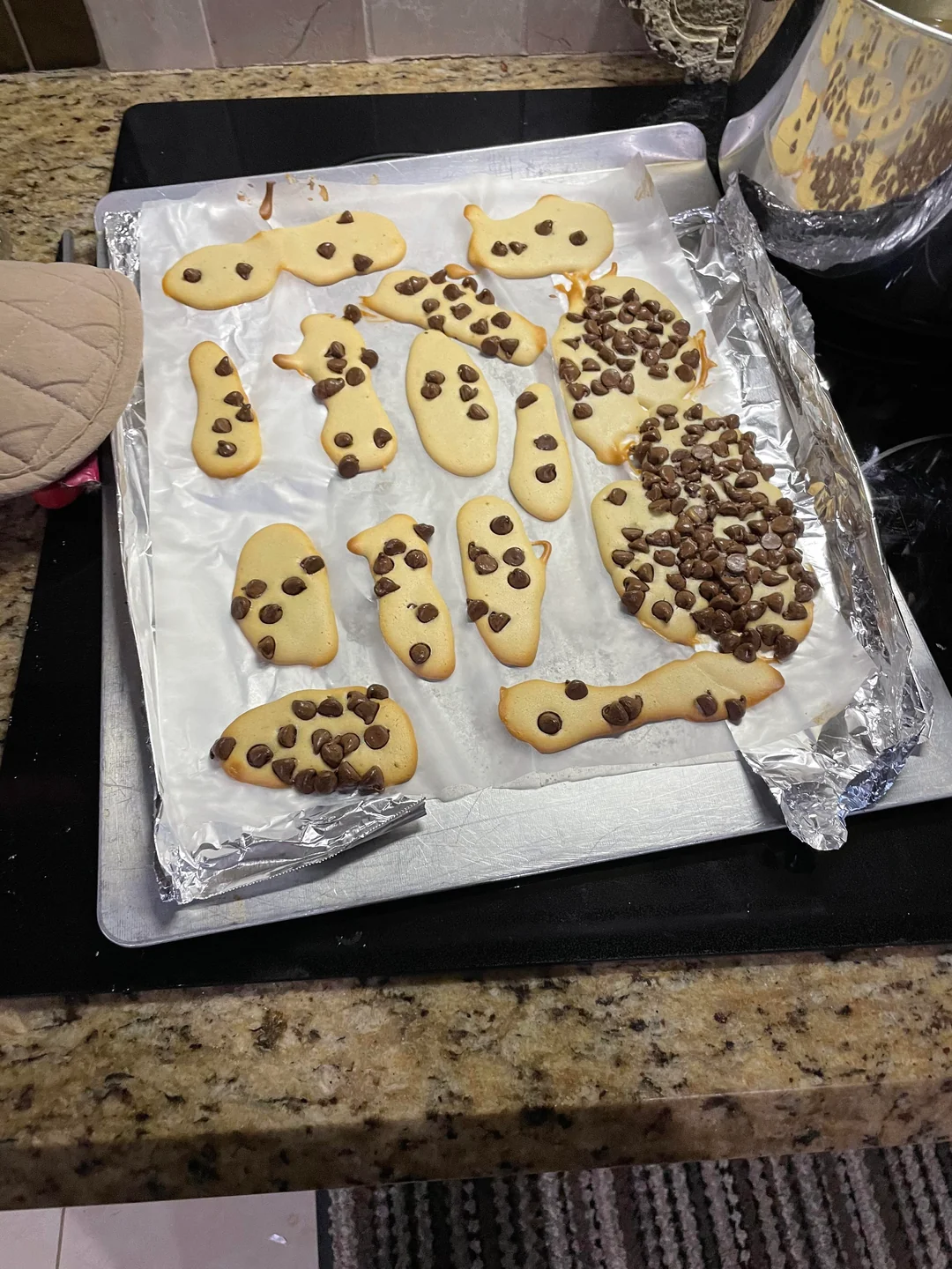 A baking tray with various homemade cookies topped with chocolate chips, some irregularly shaped, on a foil-lined sheet in a kitchen setting.