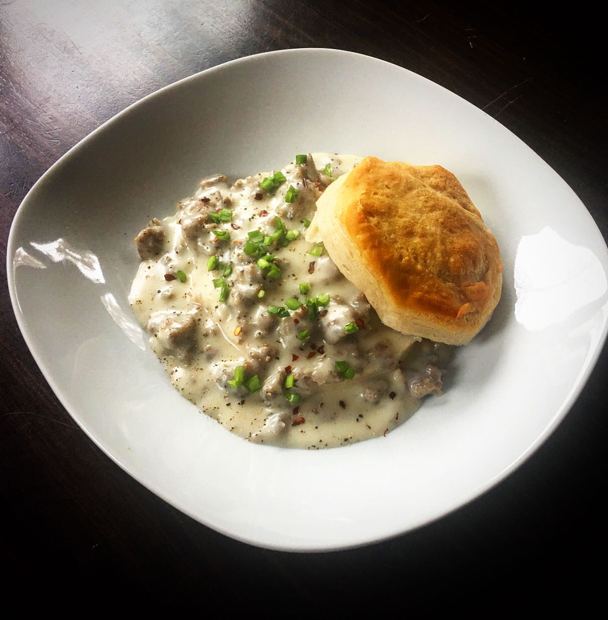 A golden biscuit sits on creamy sausage gravy, garnished with chopped green herbs, served on a white plate over a dark wooden table.