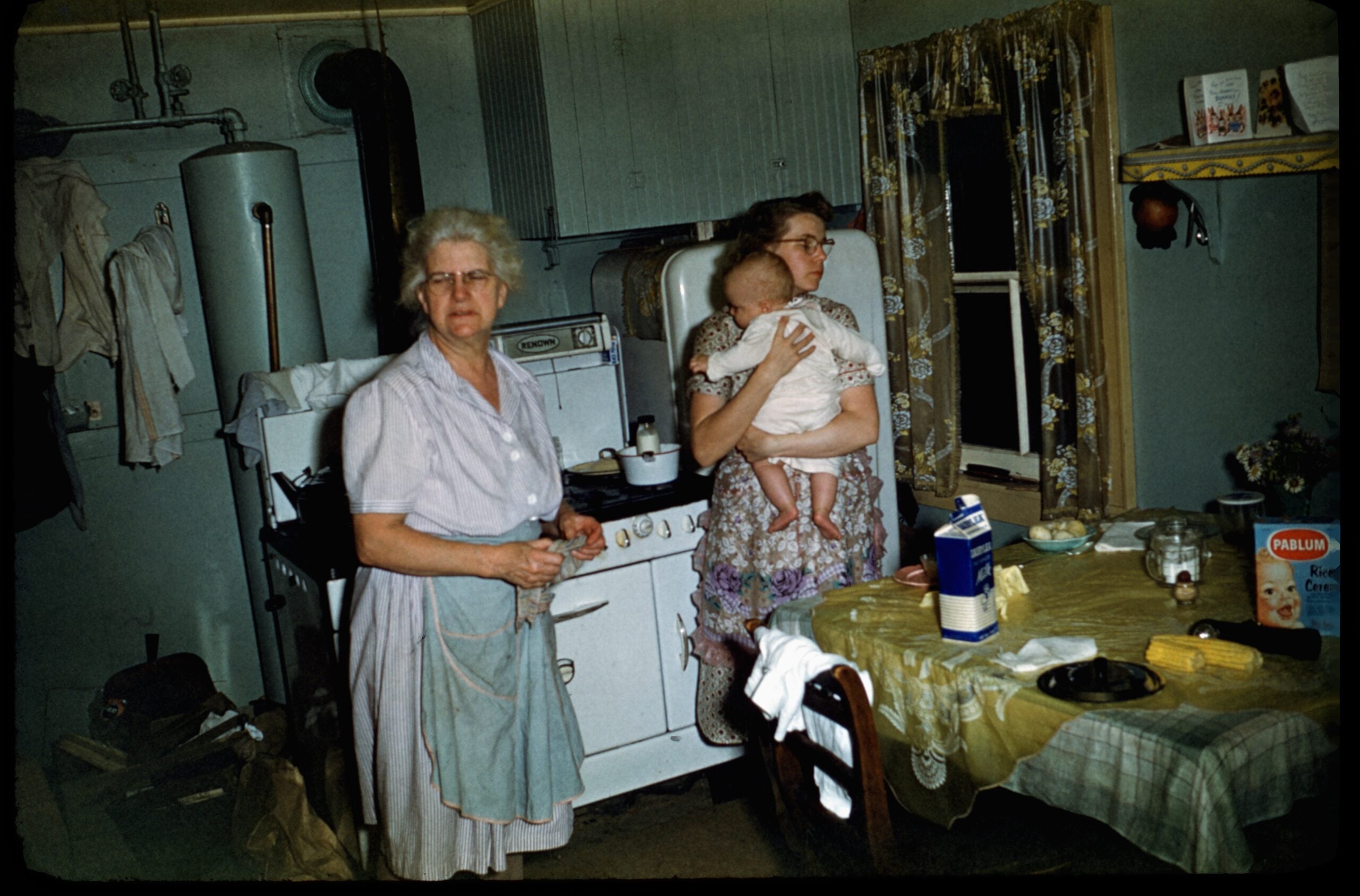 Two women in an old-fashioned kitchen: one stands by the stove in an apron, while the other, seated, holds a baby. The table is set for a meal, with groceries and dishes visible. The room has patterned curtains and vintage appliances.