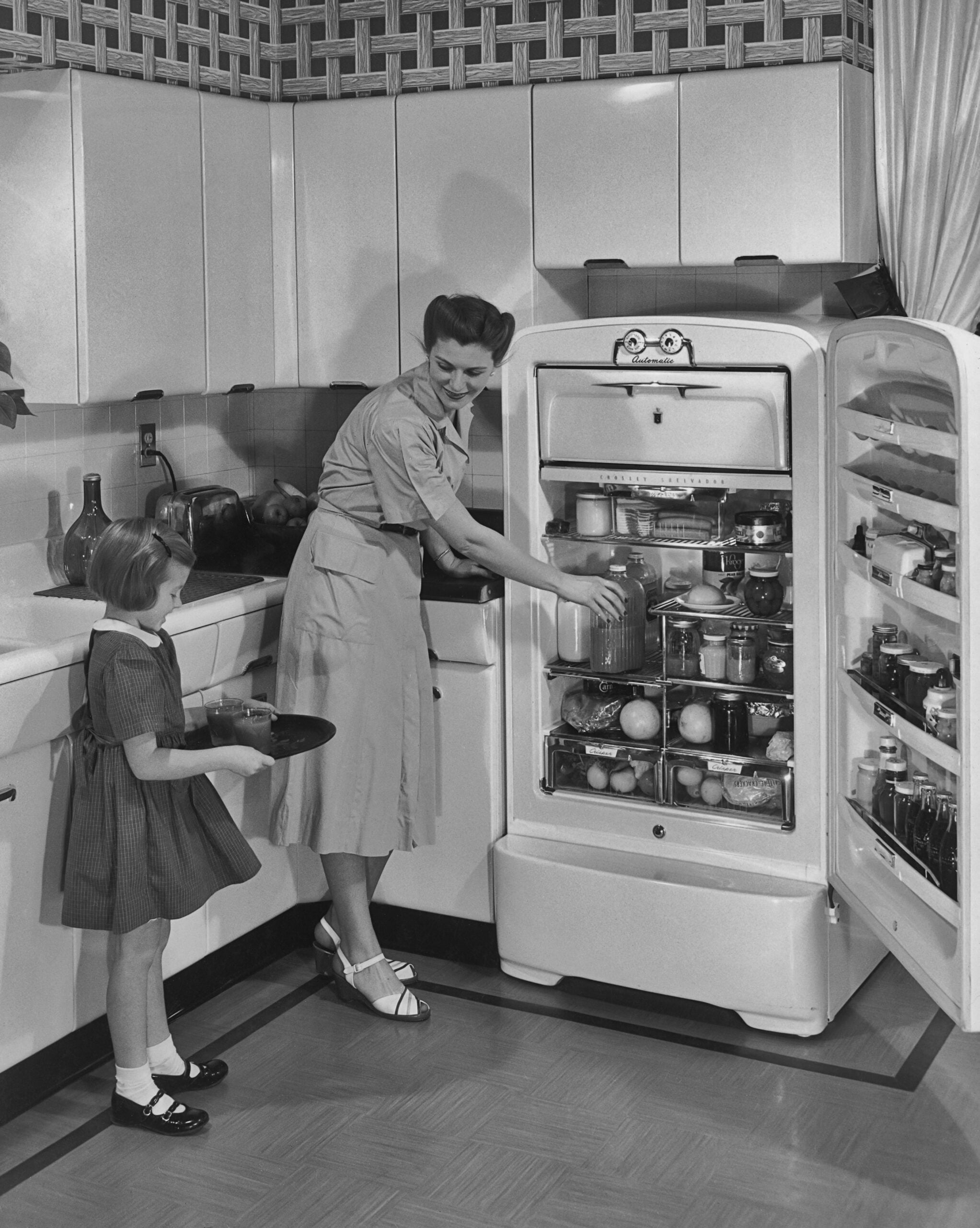 A woman and a young girl stand in a vintage kitchen. The woman is arranging food in an open, fully stocked refrigerator while the girl holds a plate and looks on. The scene is in black and white.