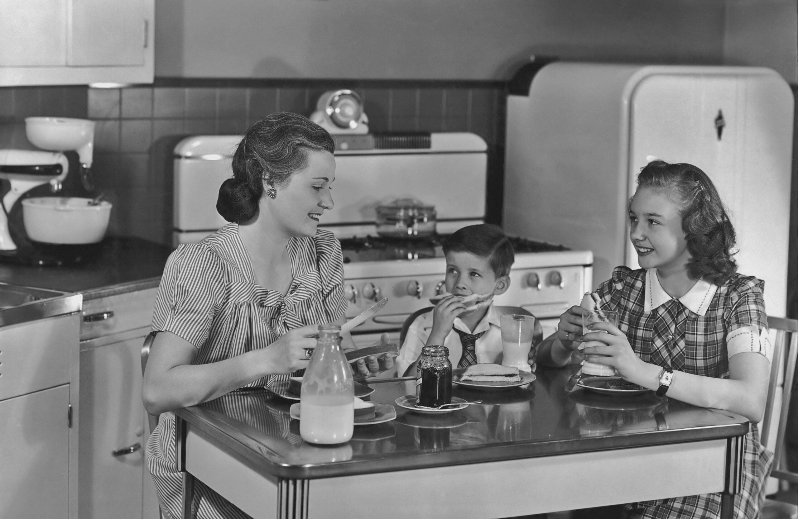 A woman, young girl, and boy sit at a kitchen table, eating breakfast. The kitchen has vintage appliances, and milk, bread, and jam are on the table. The scene appears to be from the 1950s.