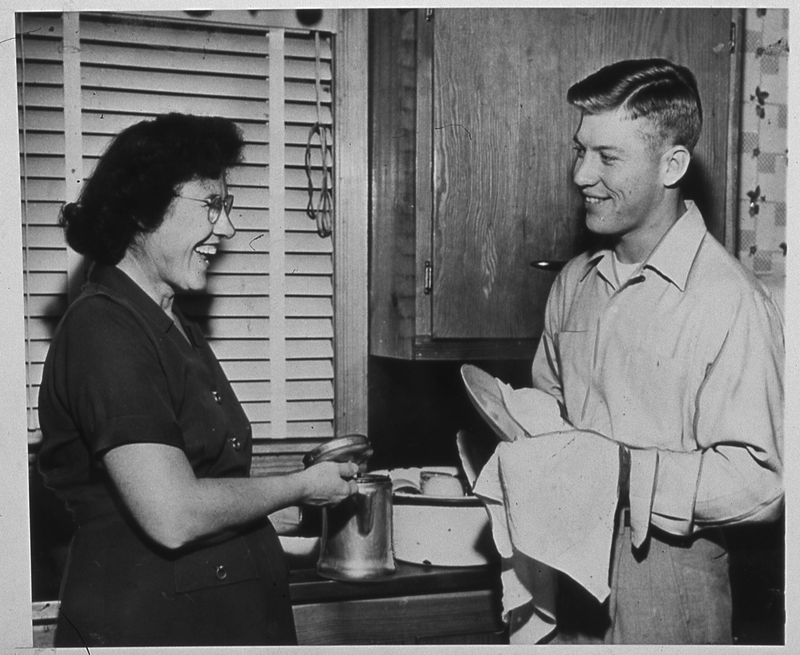A woman and a young man smile at each other in a kitchen. The woman washes dishes while the man dries a plate with a towel.