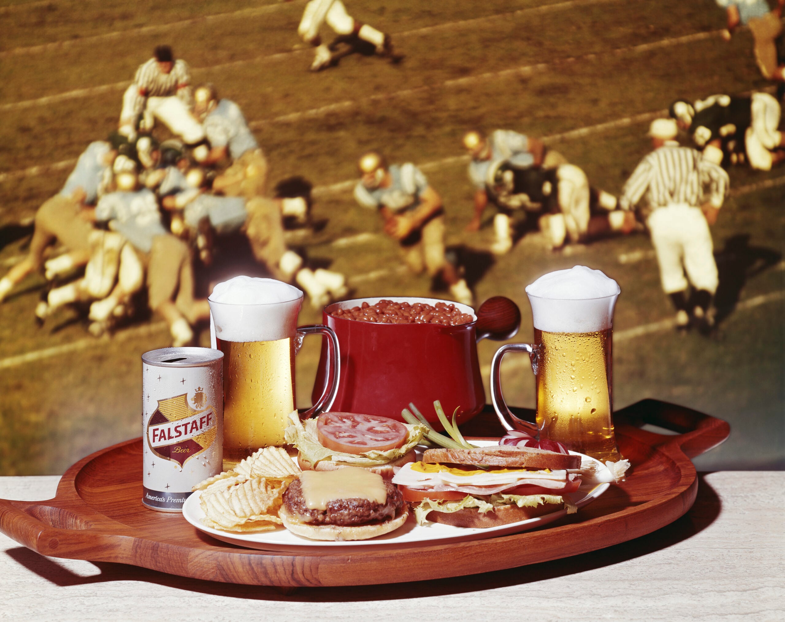 A tray with burgers, hot dogs, chips, onion rings, a can of Falstaff beer, two mugs of beer, and a bowl of chili, with a vintage football game showing in the background.