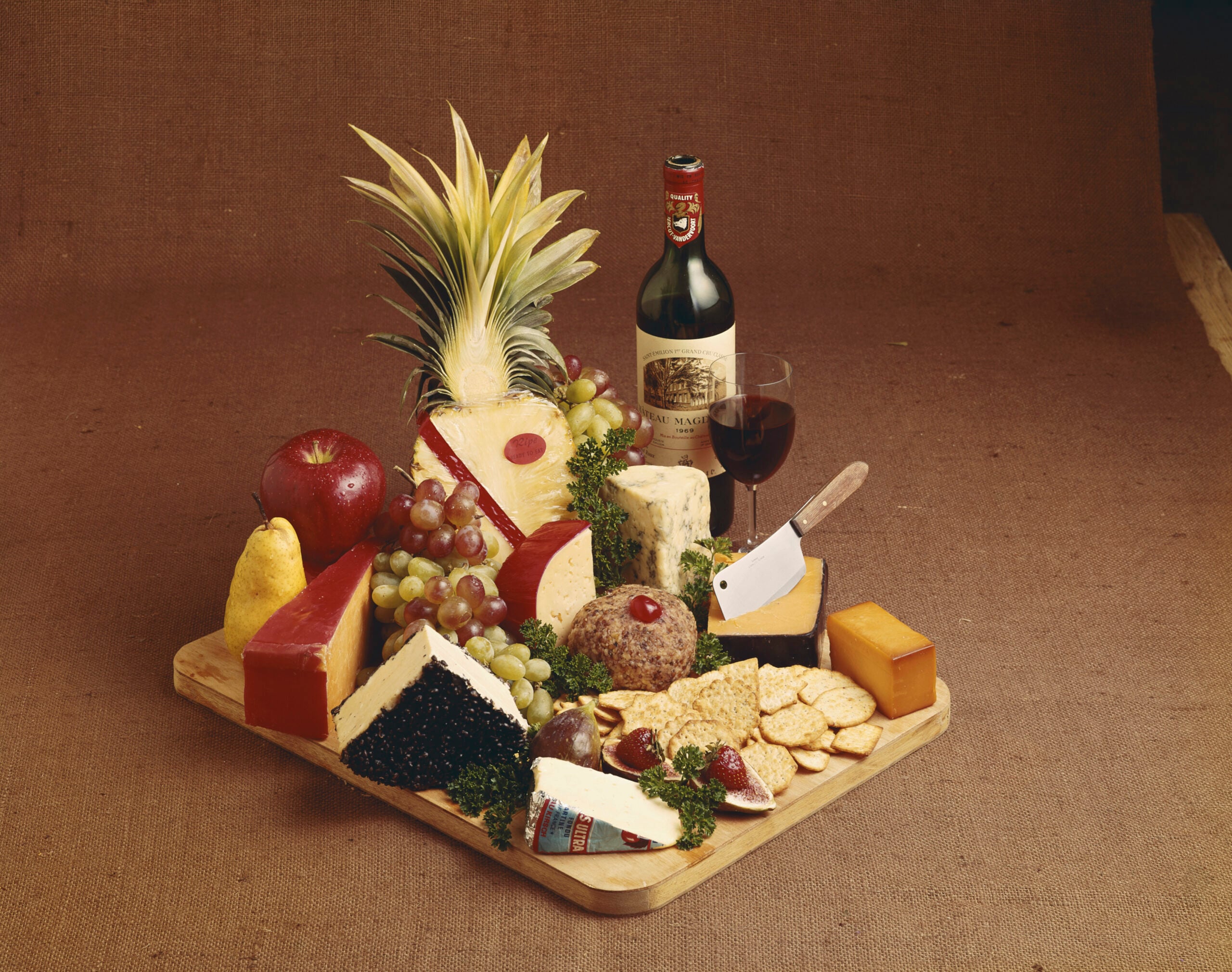 A wooden board with assorted cheeses, crackers, fruit, a wine bottle, and a glass of red wine. A pineapple and grape bunches decorate the arrangement, along with a small cheese knife and parsley garnish.