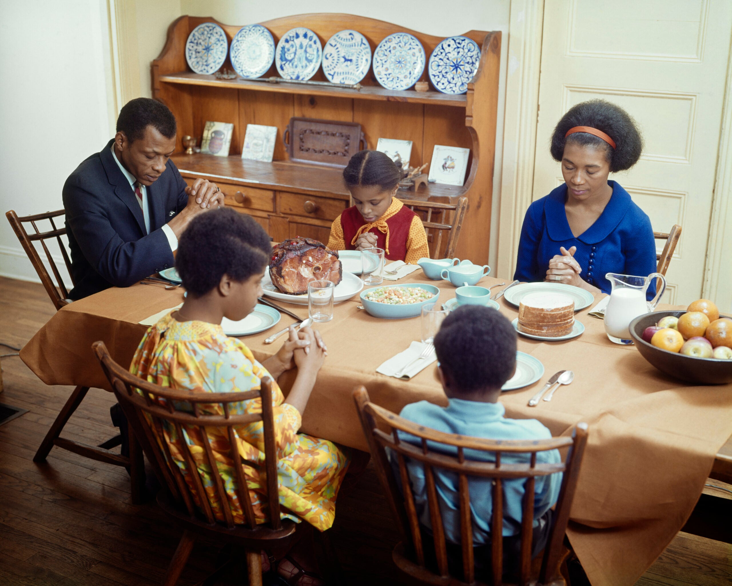 A family of five sits at a dining table with food, heads bowed and hands clasped in prayer. The table is set with dishes, and a wooden hutch with plates is in the background.
