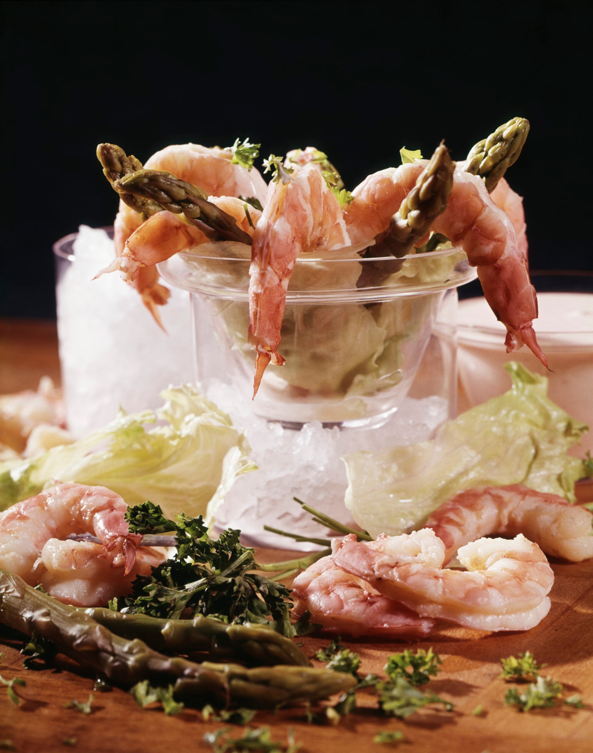 Shrimp and asparagus are arranged in a glass dish over ice, surrounded by lettuce, parsley, and more shrimp, with a small container of dipping sauce in the background.