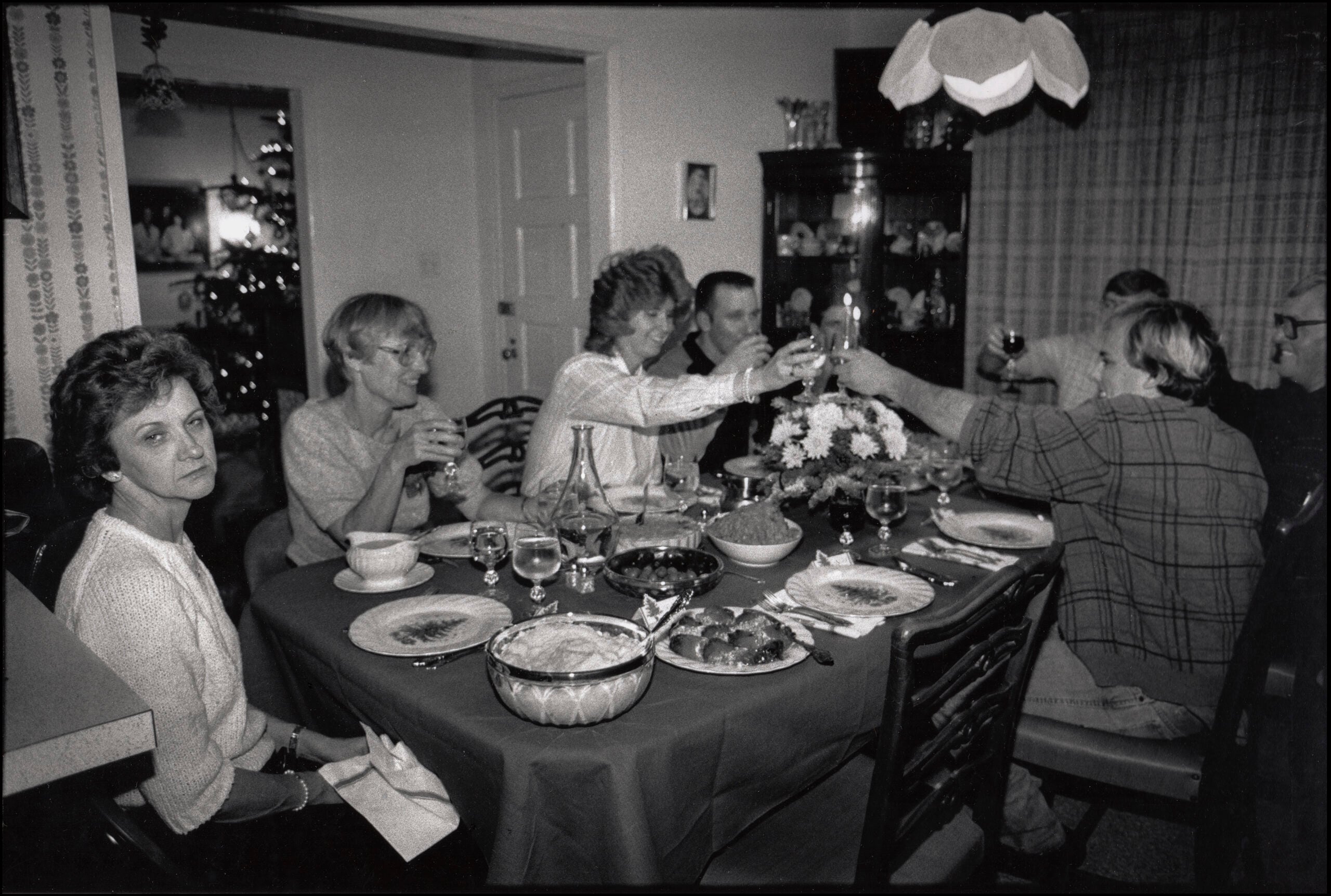 1980s family life. Seven people sit around a dining table set with food and drinks; six toast each other, smiling, while one woman on the left looks away from the group with a neutral expression. The room is warmly lit and decorated.