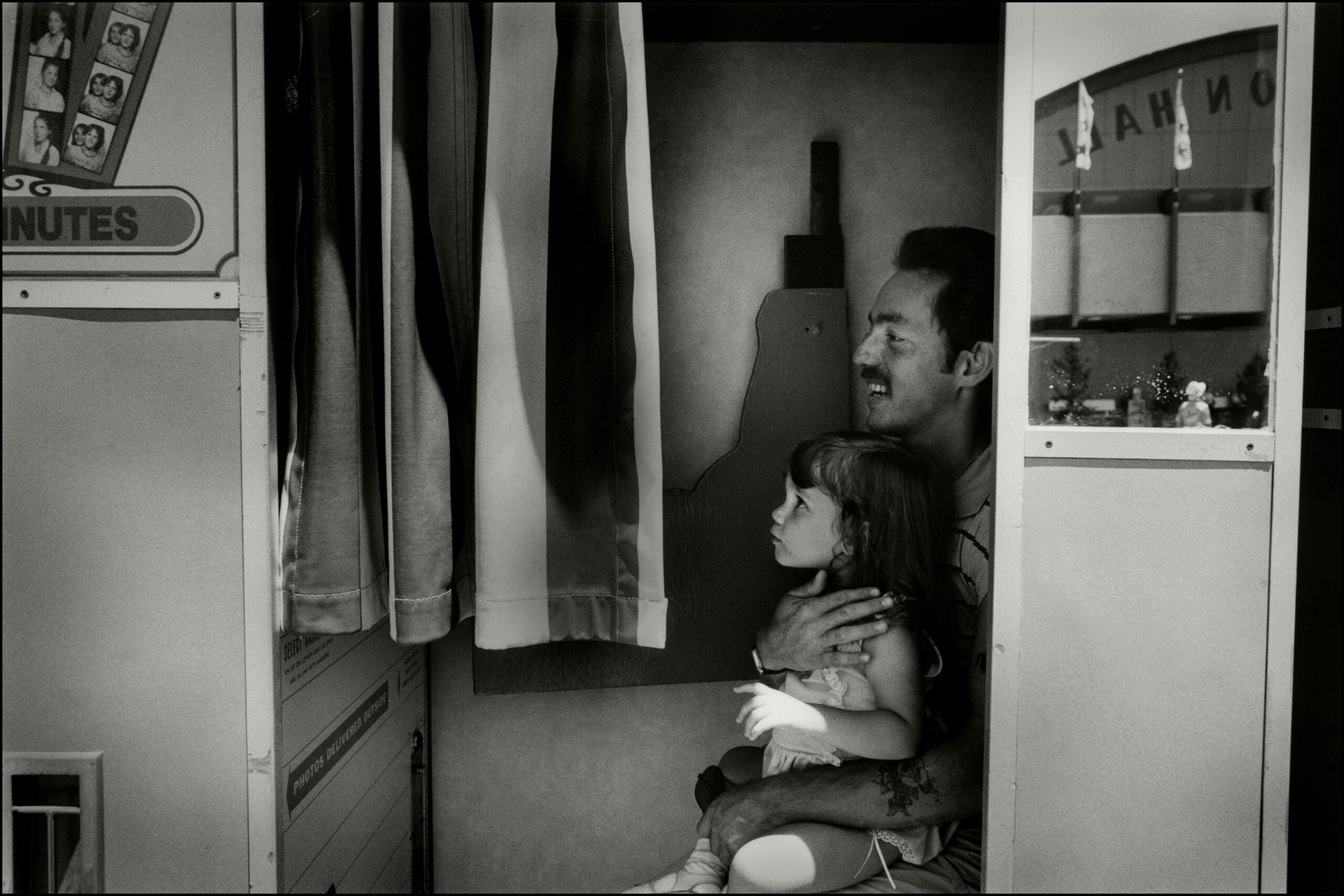 A man hugs a young girl on his lap inside a photo booth, both looking out as light filters through the curtain. Small toy decorations are visible on the booth’s shelf.