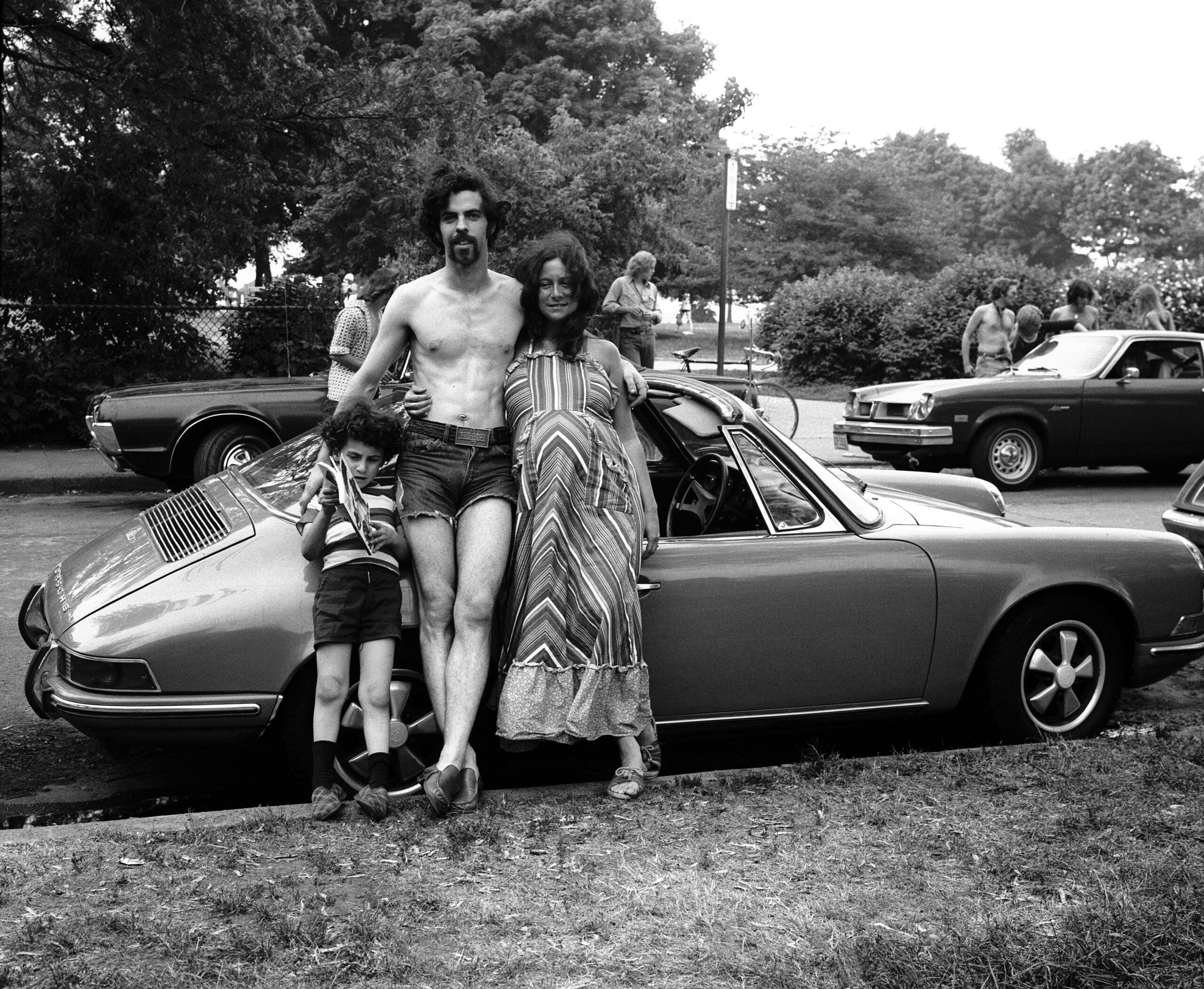 A man, woman, and child stand by a classic car parked on a street. The man is shirtless, the woman is smiling, and the child leans against them. Other people and cars are visible in the background.