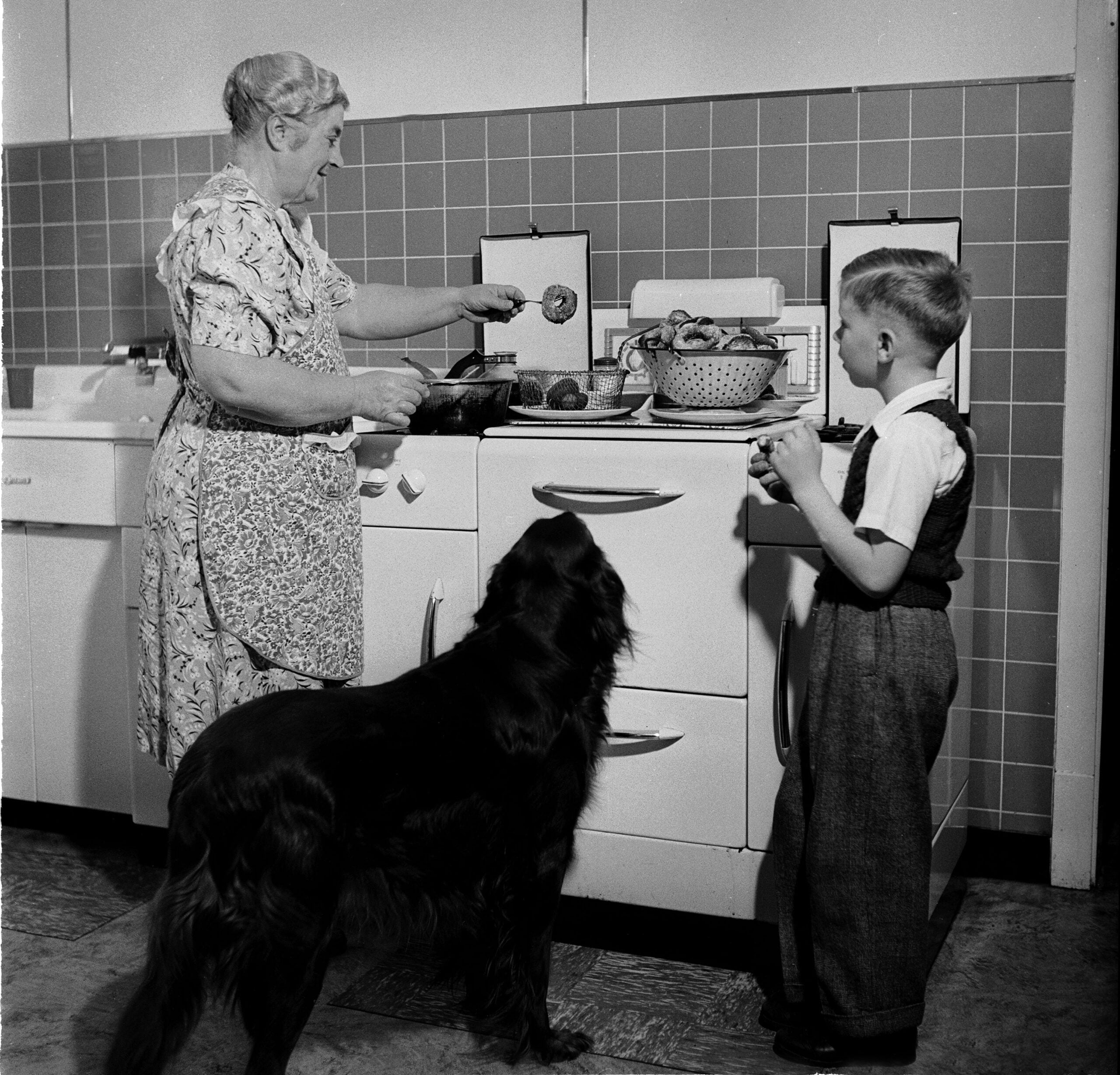 An older woman cooks at a stove while a young boy and a large black dog watch her in a kitchen with tiled walls and retro appliances.