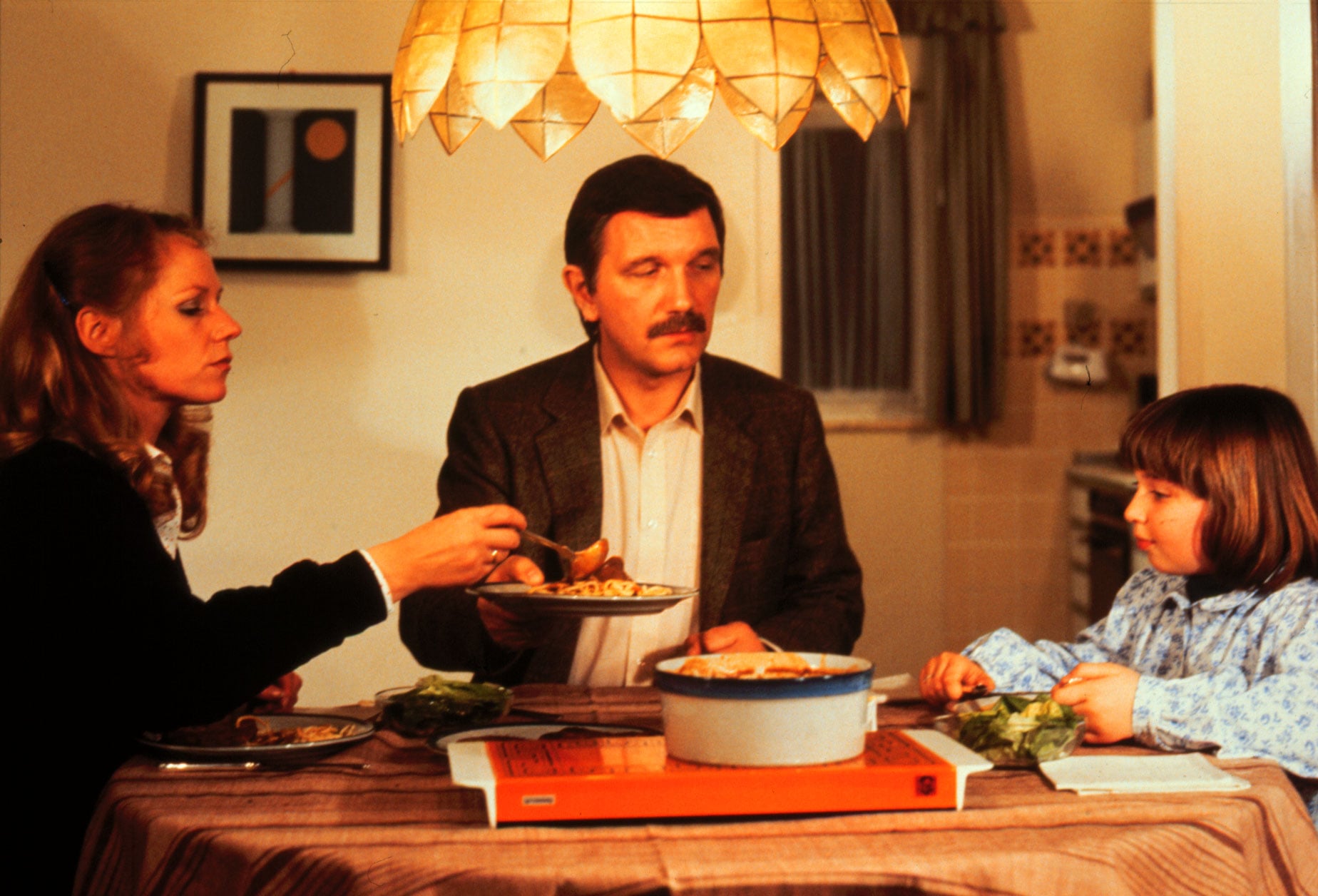 A woman, man, and child sit at a dining table with plates of food, sharing a meal under a decorative light. A framed picture and kitchen are visible in the background.