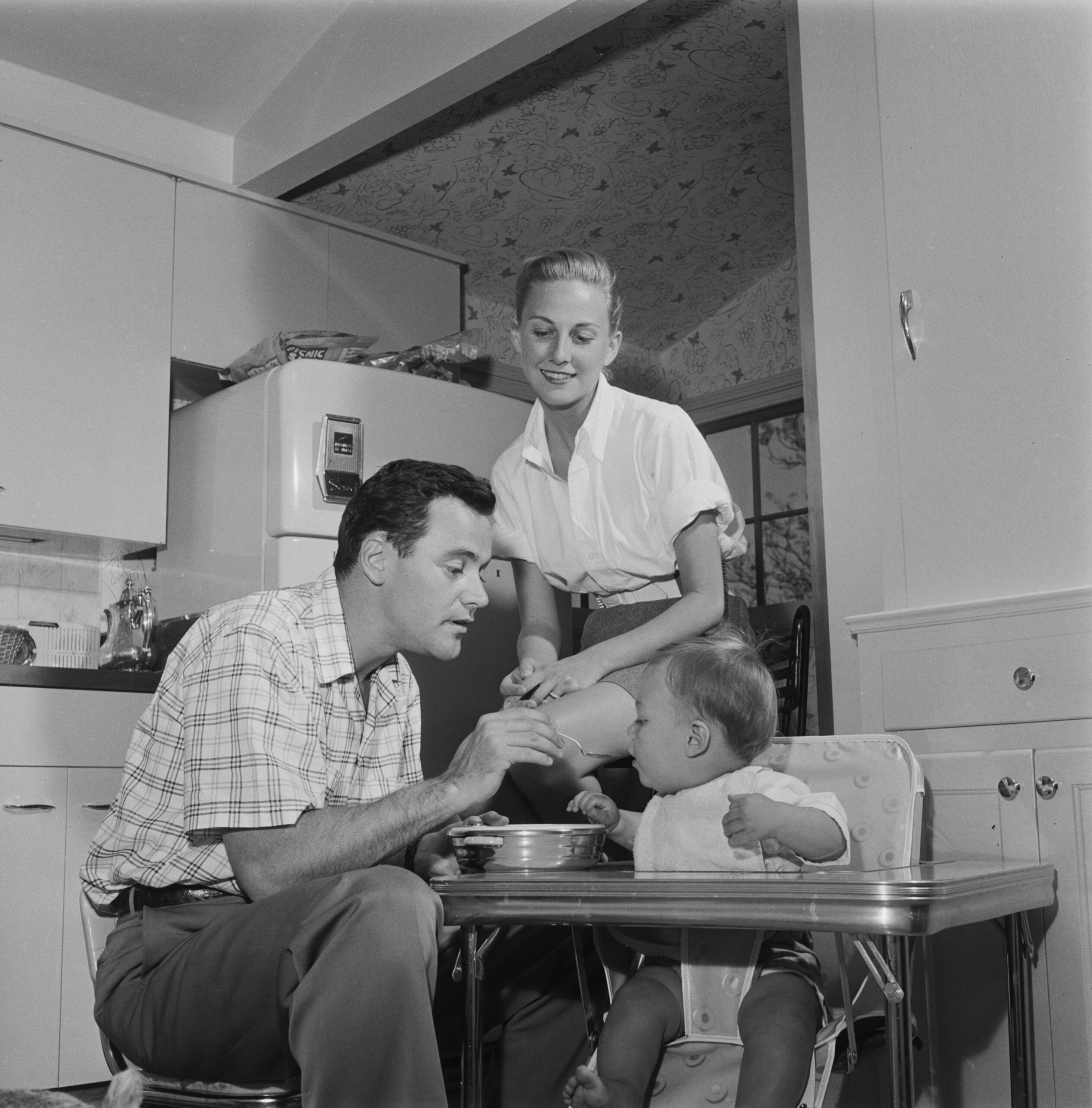 A man feeds a baby in a high chair while a woman smiles and leans over in a vintage kitchen setting.