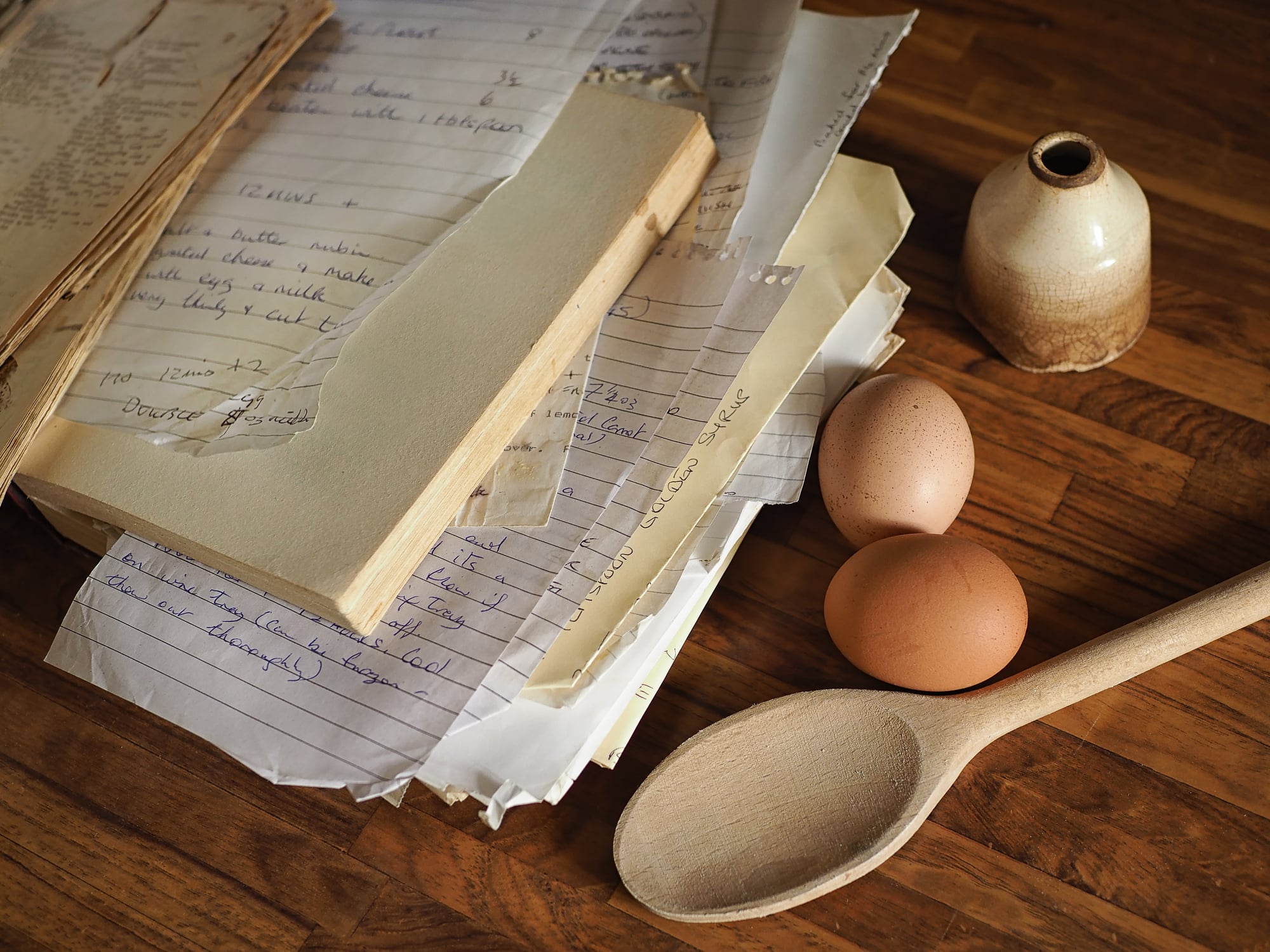 A stack of handwritten recipes, a wooden spoon, three eggs, and a small brown ceramic pot—classic kitchen items—are arranged on a wooden surface.