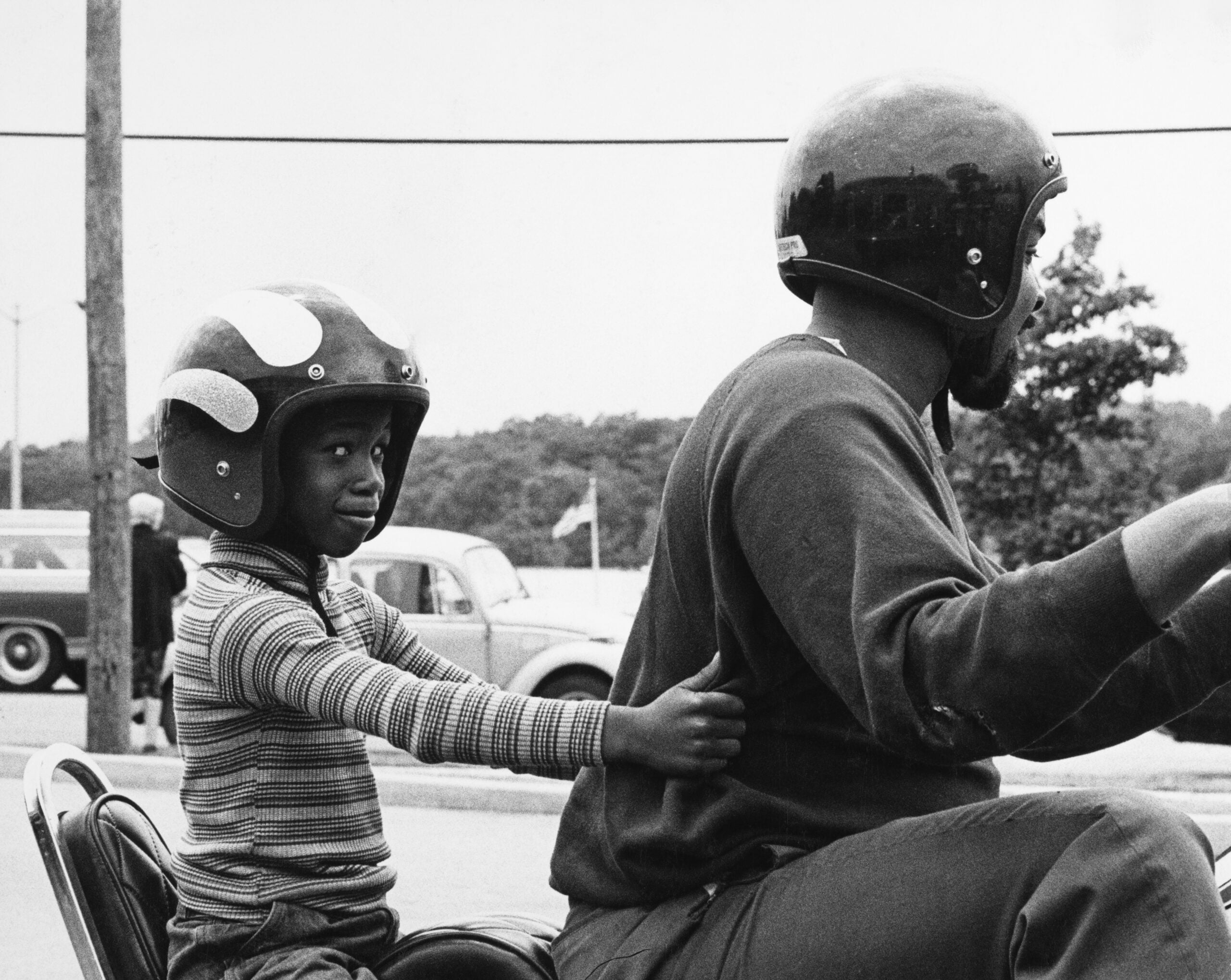 A child in a striped shirt and helmet sits behind an adult on a motorcycle, holding on and smiling. Both wear helmets, and cars and trees are visible in the background.