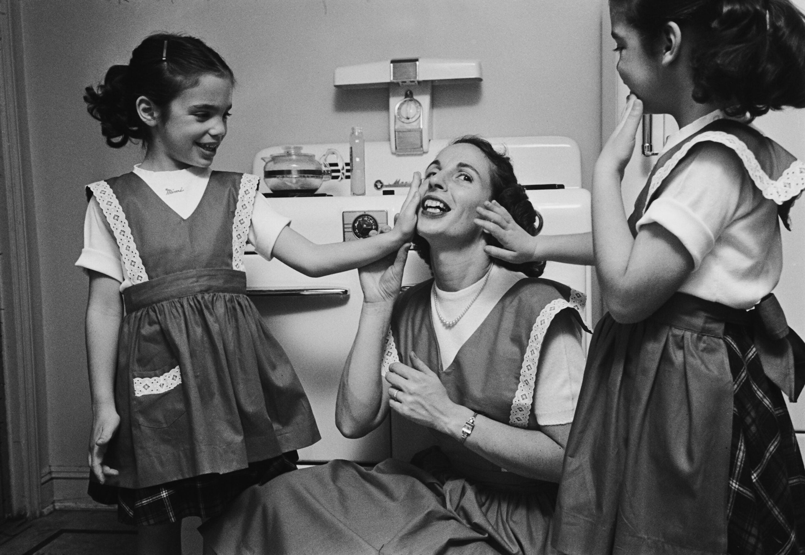 A woman kneels between two young girls in matching dresses. The girls are smiling and playfully pulling at the woman's cheeks in a kitchen setting. The atmosphere appears joyful and lighthearted.