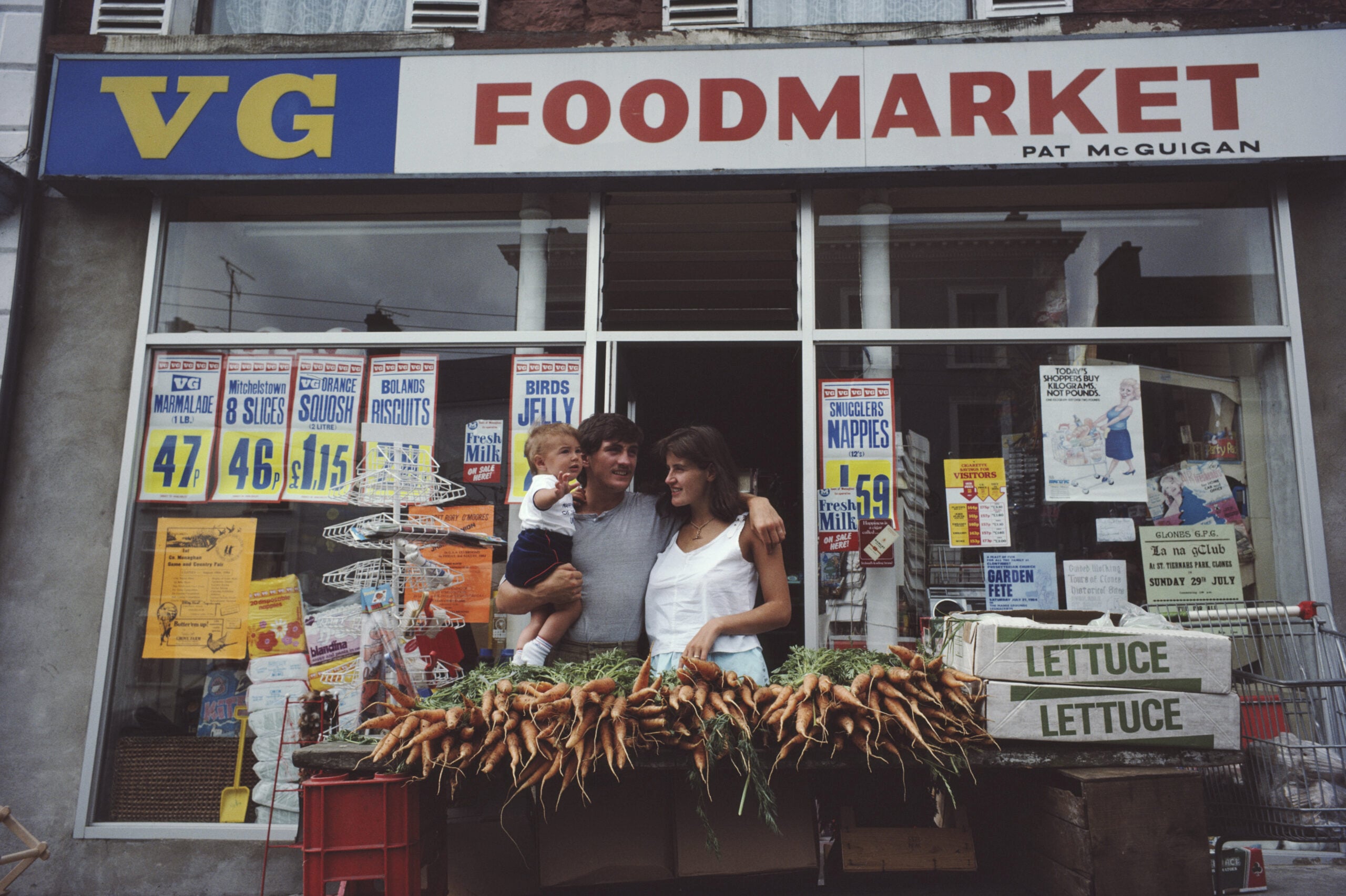 A family stands in front of a food market, holding a child, with crates of fresh carrots and two signs labeled “LETTUCE” on display. Posters with prices are visible in the store windows above them.